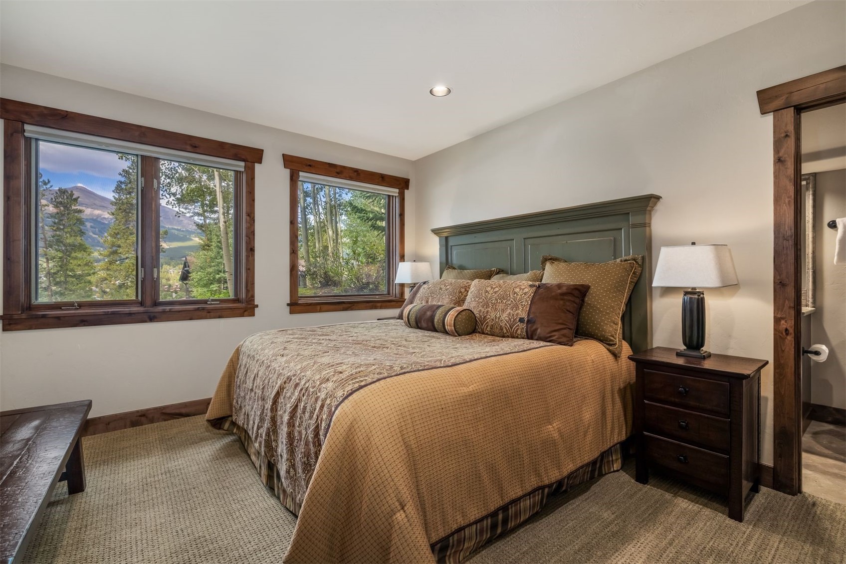 This cozy bedroom features a queen-sized bed with a dark green headboard and warm-toned, patterned bedding. Two large windows with rustic wood trim frame a scenic view of the surrounding trees and mountains, while a dark wood nightstand with a lamp sits to the right. The room is finished with neutral walls and textured carpeting, creating a comfortable and inviting mountain-style retreat.