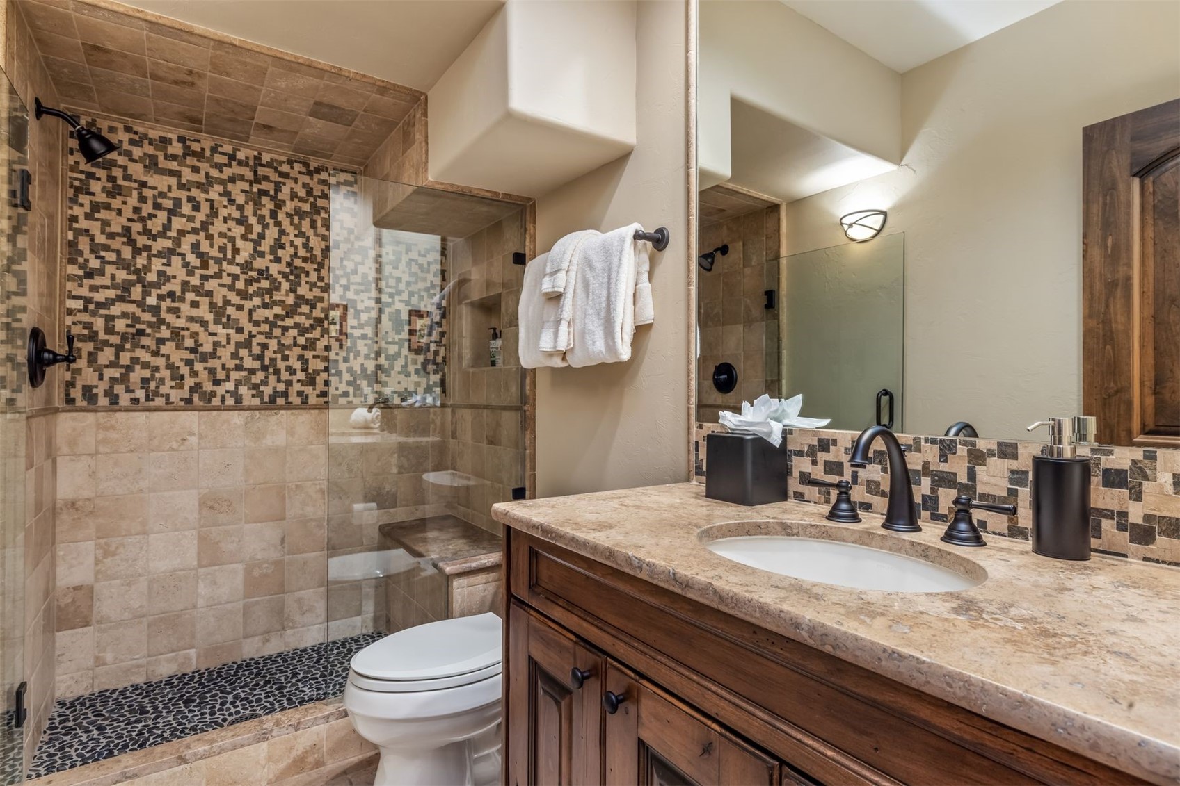 This bathroom features a warm, rustic aesthetic with a stone-topped vanity and dark wood cabinetry. A walk-in shower is enclosed by a glass panel and showcases a combination of neutral stone tiles and a decorative mosaic accent wall. The space is completed by a toilet positioned next to the shower and a wall-mounted mirror reflecting the vanity area.