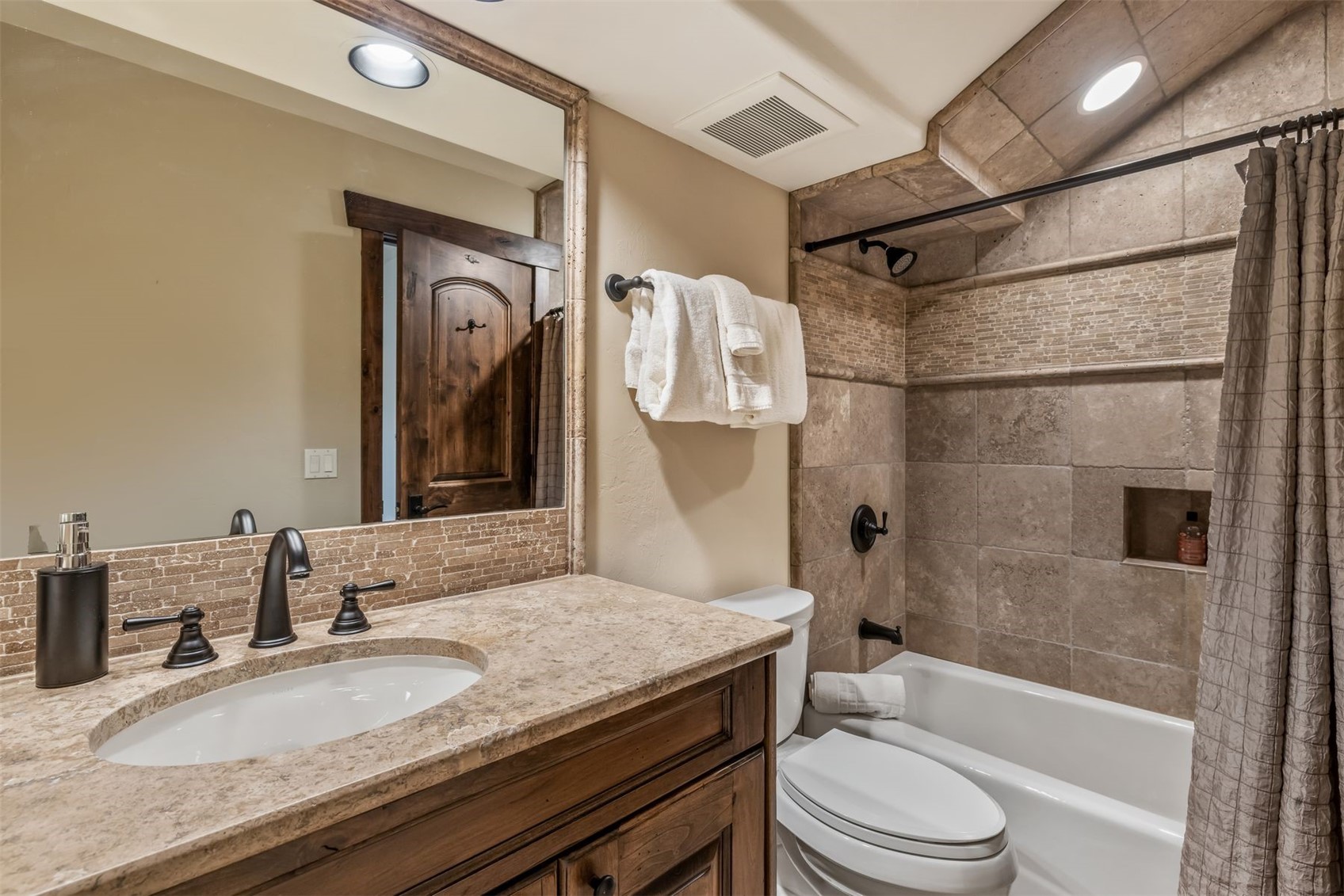 This compact bathroom features a rustic aesthetic with a stone-topped vanity, dark bronze fixtures, and a tiled tub-shower combination. The space is accented by a decorative mosaic tile backsplash and a framed mirror, creating a warm and cohesive look. The perspective is a medium-wide shot capturing the vanity, toilet, and shower area in a single view.