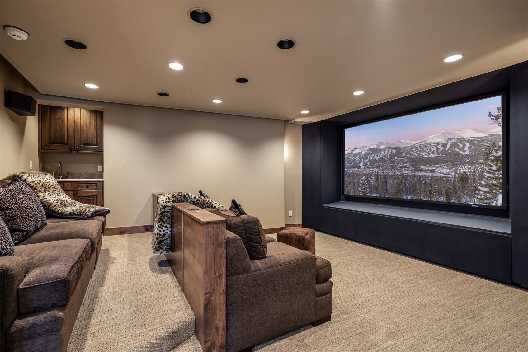 This cozy home theater features tiered seating with plush brown sofas and a large, wall-mounted screen displaying a scenic mountain landscape. To the side, a small wet bar area with wooden cabinetry and a sink adds convenience to the space. The room is finished with neutral carpeting and recessed lighting, creating an inviting atmosphere for entertainment.