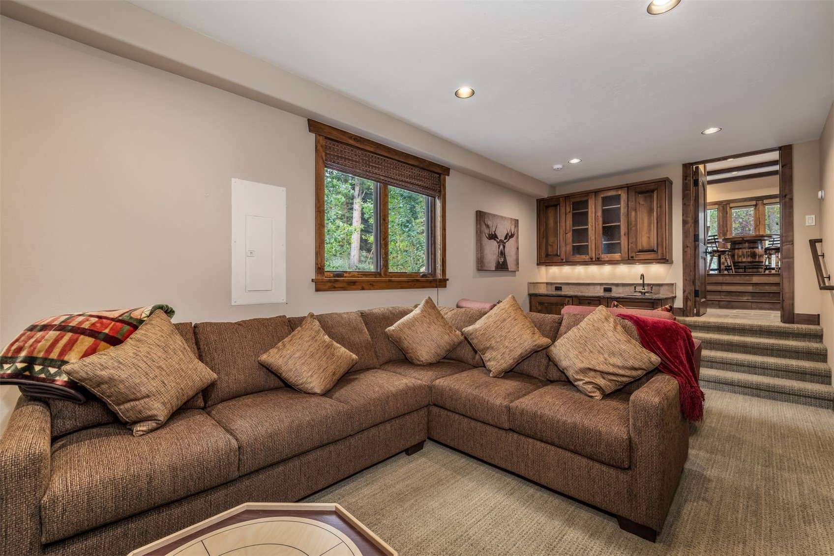This cozy living room features a large, L-shaped brown sectional sofa adorned with patterned throw pillows and a plaid blanket. The space includes a wet bar area with wooden cabinetry and a small sink, while a window provides a view of the surrounding trees. A set of carpeted stairs leads up to an adjacent room, creating a multi-level layout that feels warm and inviting.