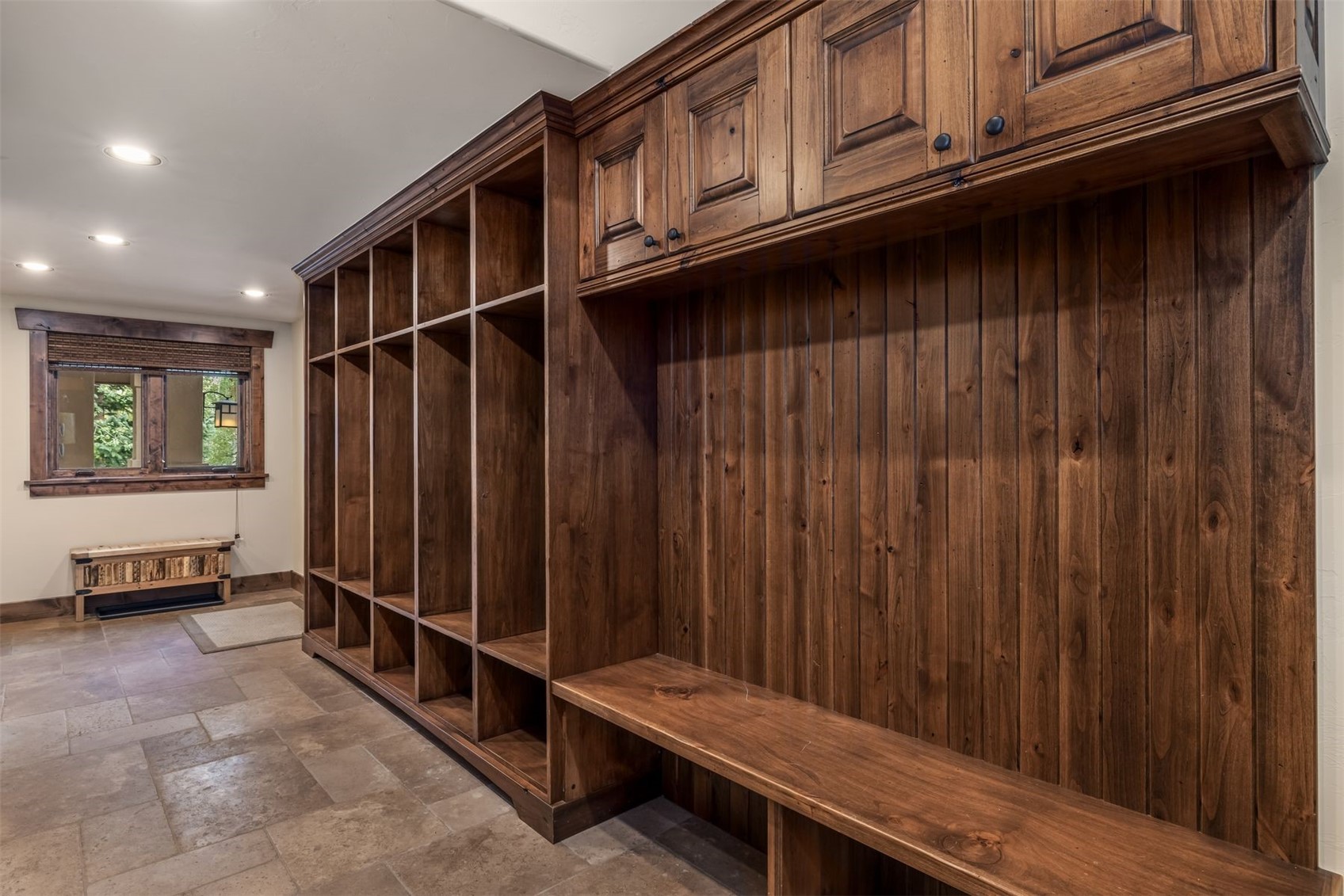 This mudroom area features extensive custom wood cabinetry with built-in cubbies and a long bench seat, providing ample storage and organization space. The room is finished with neutral stone tile flooring and warm wood trim, creating a rustic and functional aesthetic. A window at the end of the hall allows natural light to enter, highlighting the rich wood tones and clean lines of the built-in storage.