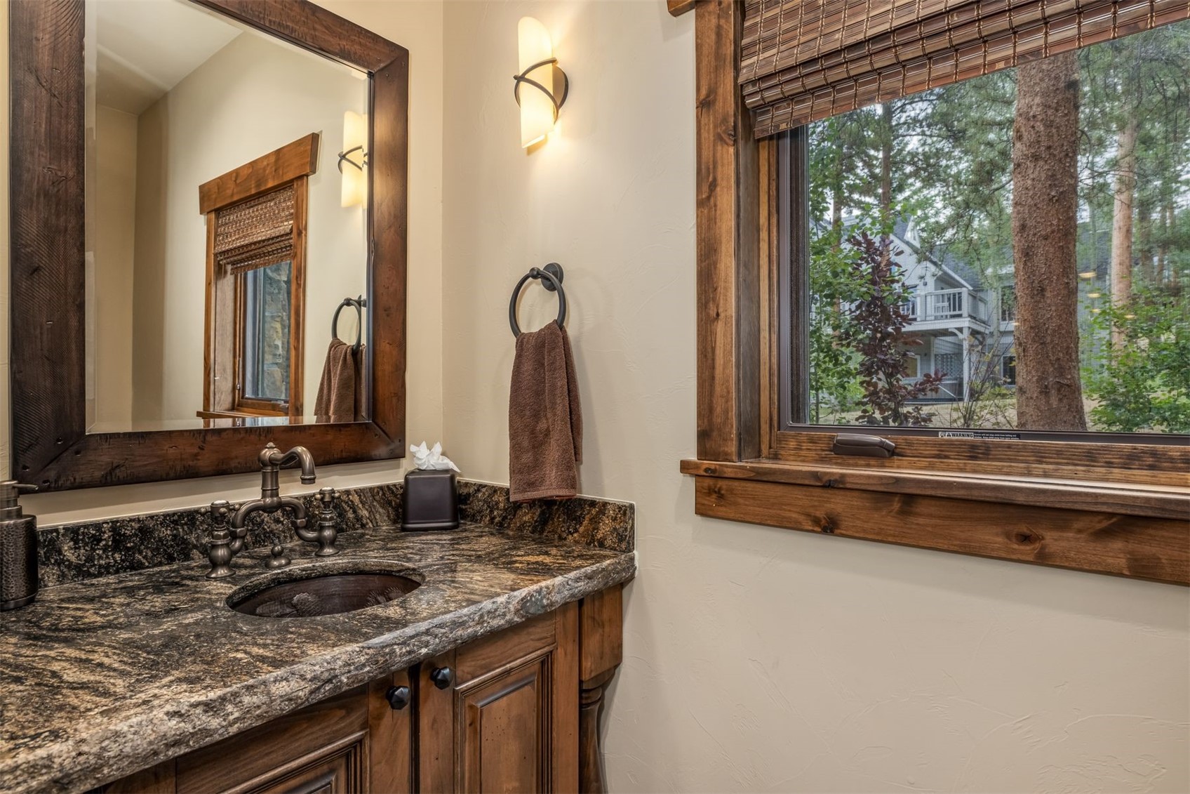 This rustic-style powder room features a dark, patterned stone countertop with an integrated sink and a dark wood vanity. A large mirror with a matching wood frame hangs above the vanity, reflecting a window with a woven shade, while a wall-mounted light fixture provides warm illumination. The space is finished with neutral-toned walls and a window that looks out onto a wooded exterior view.