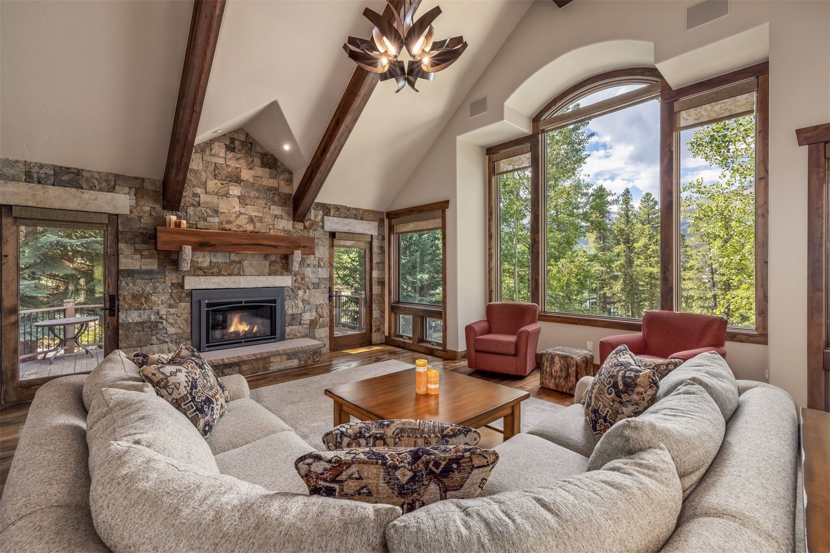 This spacious living room features a vaulted ceiling with exposed timber beams and a striking, multi-armed chandelier. A prominent stone-clad chimney breast serves as the focal point, complemented by a large, light-colored sectional sofa and two red armchairs. Expansive windows provide abundant natural light and scenic views of the surrounding forest, creating an inviting and rustic atmosphere.