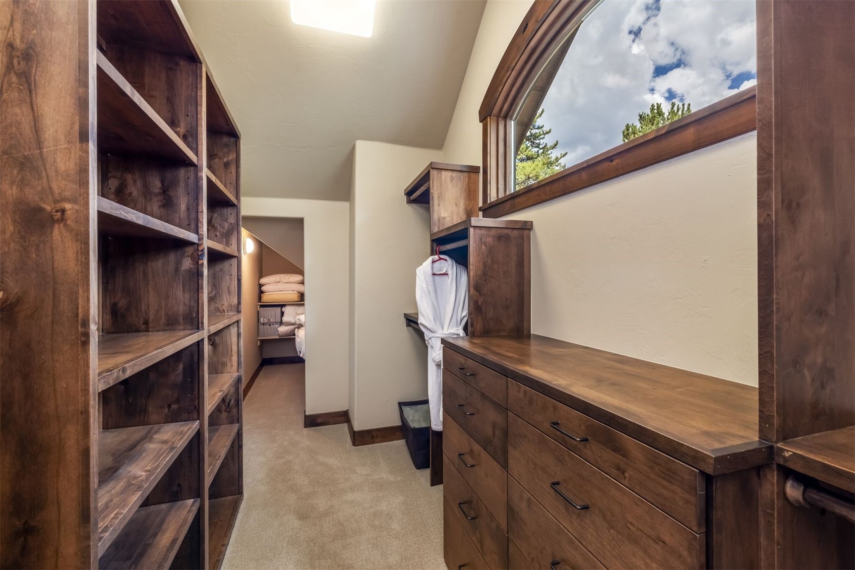 This spacious walk-in closet features custom dark wood cabinetry, including open shelving units and a built-in dresser with multiple drawers. A white robe hangs in a dedicated alcove, while a large arched window allows natural light to brighten the room. The space leads into a secondary storage area, showcasing a well-organized and functional design with neutral carpeting.