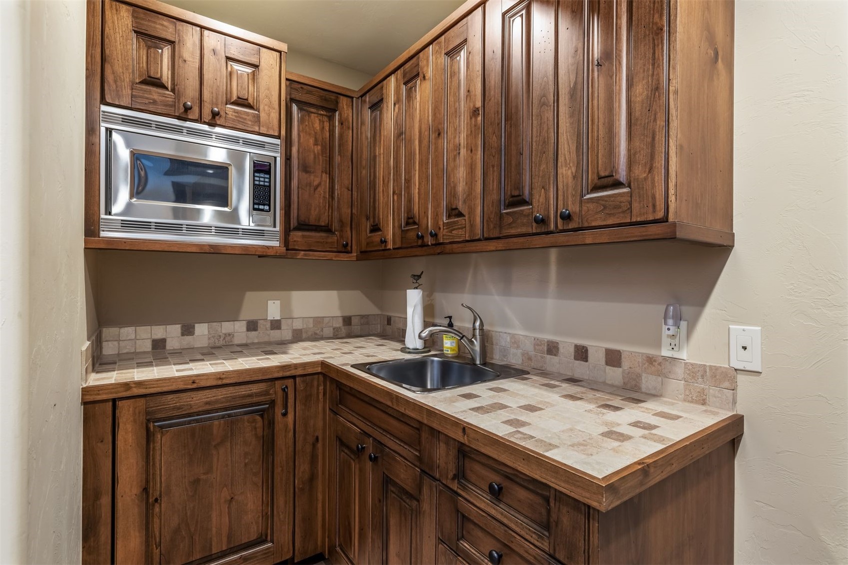 This compact kitchen area features rich, dark-toned wood cabinetry and a tiled countertop with a matching backsplash. A stainless steel microwave is built into the upper cabinets, and a small sink with a modern faucet is set into the corner workspace. The perspective is a straight-on, eye-level shot that highlights the efficient use of space and the warm, traditional aesthetic of the cabinetry.