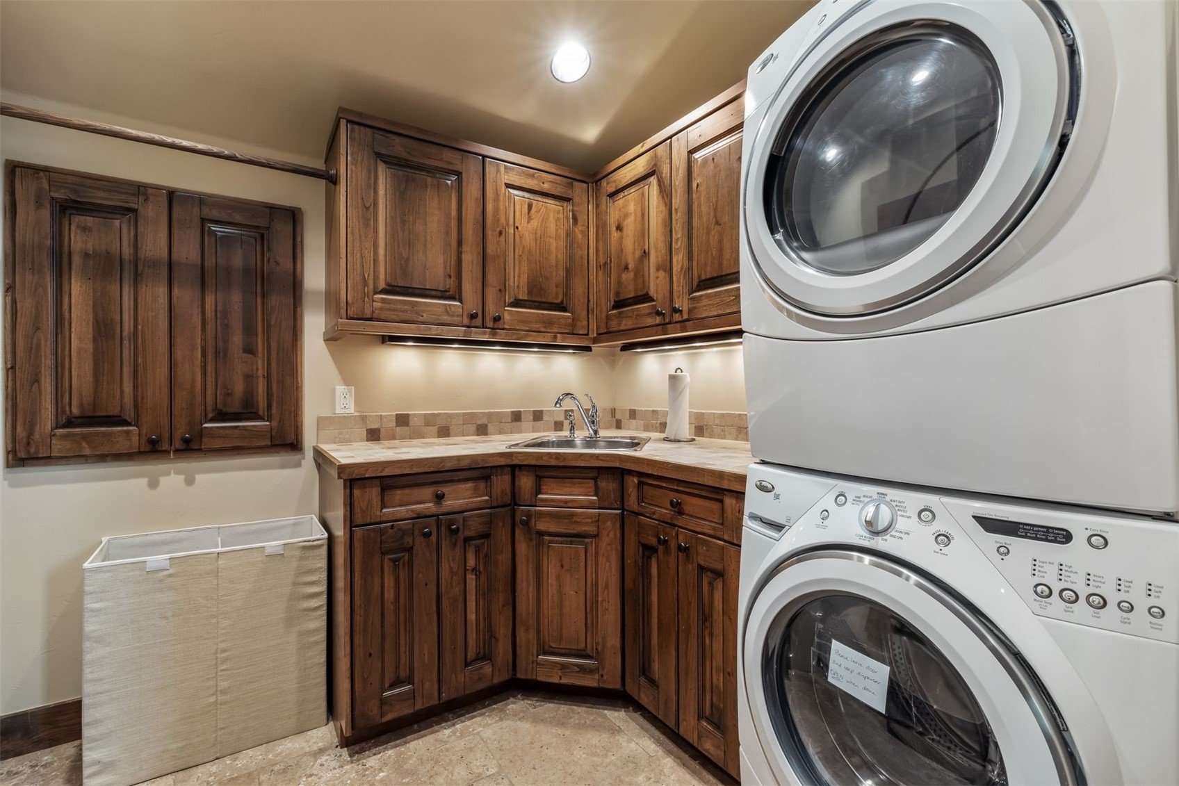 This functional laundry room features rich, dark-toned wood cabinetry providing ample storage above and below a tiled countertop with an integrated sink. A stacked washer and dryer unit stands to the right, while a fabric laundry hamper sits on the floor to the left. The space is illuminated by under-cabinet lighting and a ceiling fixture, creating a warm and organized utility area.