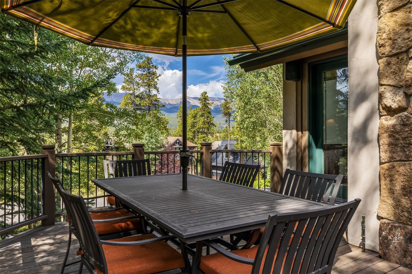 This elevated deck features a dark-toned dining set with orange cushions, shaded by a large green umbrella. The space is framed by a dark metal railing and offers expansive, scenic views of lush green trees and distant mountains under a bright, partly cloudy sky. To the right, a portion of the home's exterior is finished with textured stone cladding, creating a rustic and inviting outdoor living area.