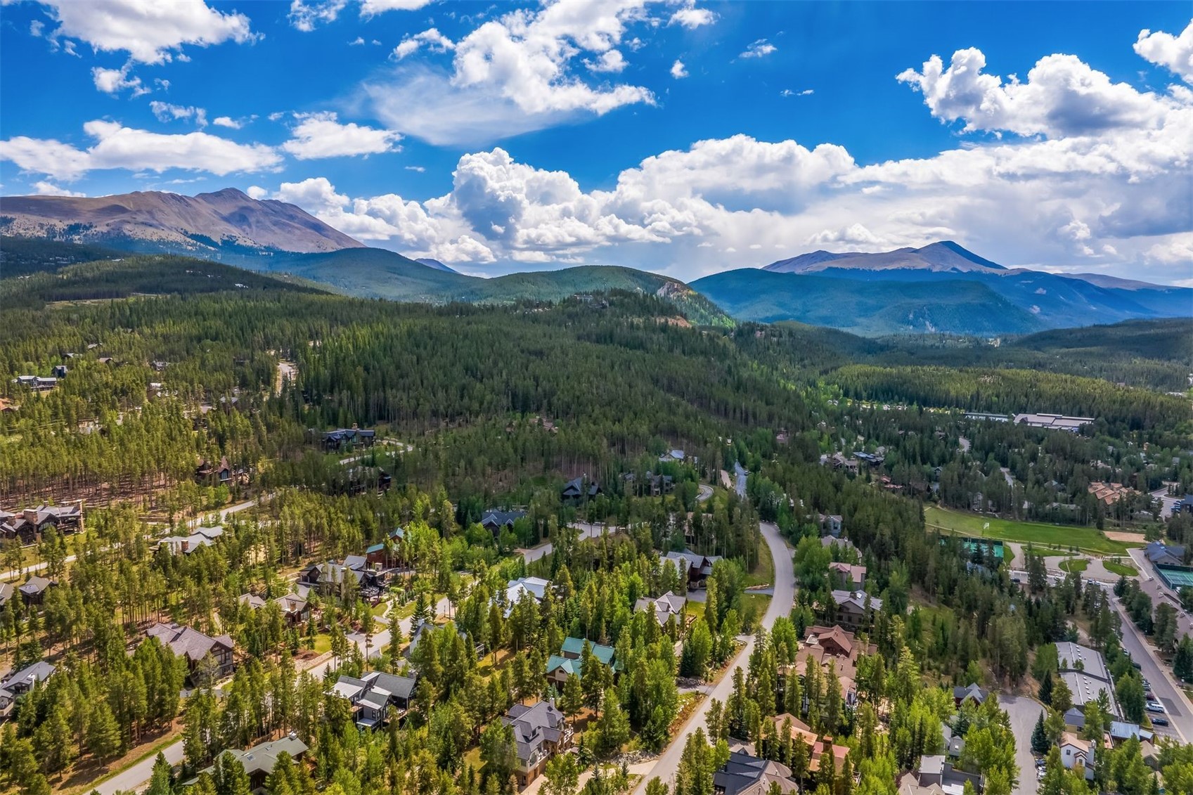 This high-angle aerial view captures a sprawling mountain community nestled within a dense evergreen forest. The landscape is dominated by rugged, snow-capped peaks under a vibrant blue sky with scattered clouds, while winding roads connect various residential homes tucked among the trees. The perspective provides a comprehensive look at the expansive natural setting and the integration of the properties into the mountainous terrain.