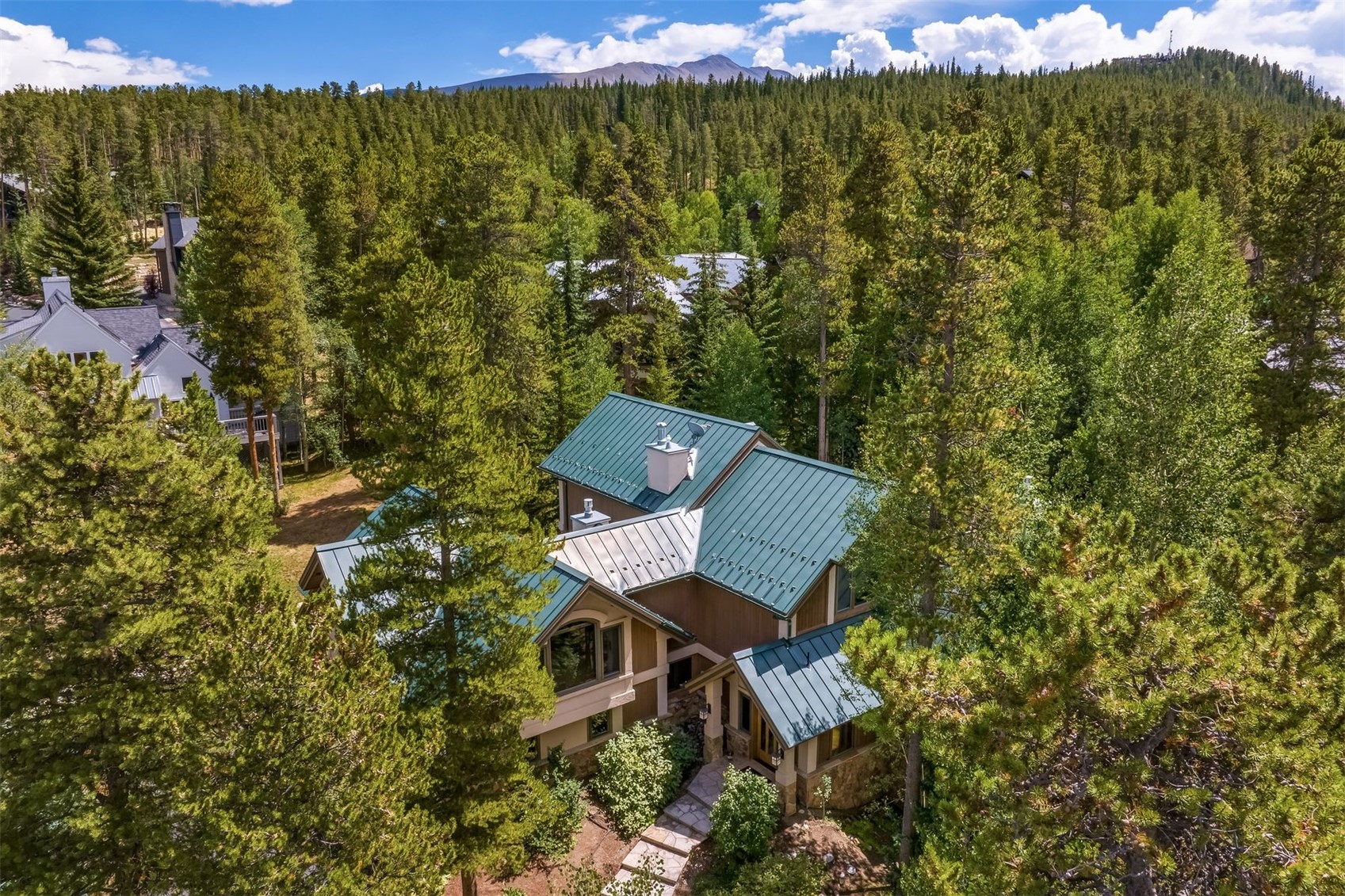 This high-angle aerial view captures a mountain-style home nestled within a dense forest of pine and aspen trees. The residence features a multi-gabled roof with green metal panels, a stone and wood-paneled exterior, and a stone walkway leading to the front entrance. In the background, a distant mountain range rises above the treeline under a bright, partly cloudy sky.