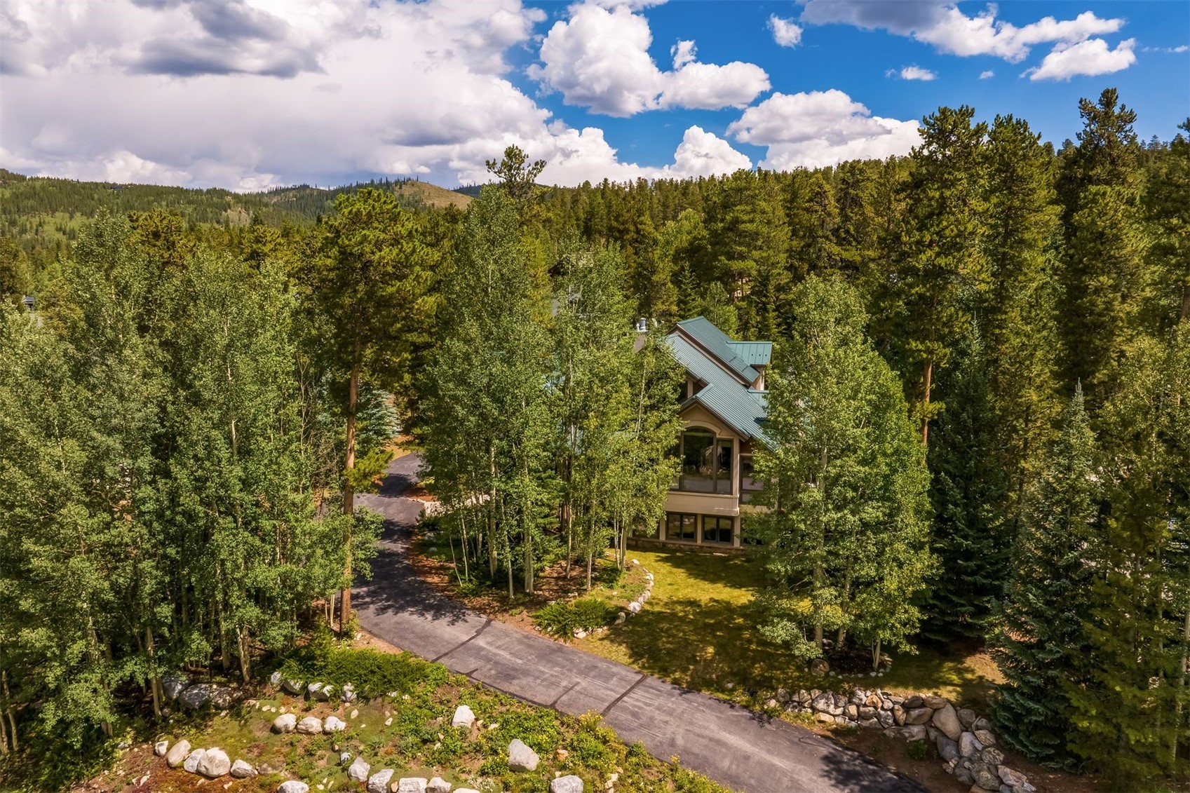 This high-angle aerial shot captures a multi-level mountain home nestled within a dense forest of pine and aspen trees. The residence features a dark green metal roof and a mix of neutral-toned siding, with a paved driveway winding through the wooded landscape toward the entrance. The setting is serene and secluded, emphasizing the property's integration into the natural, mountainous environment.