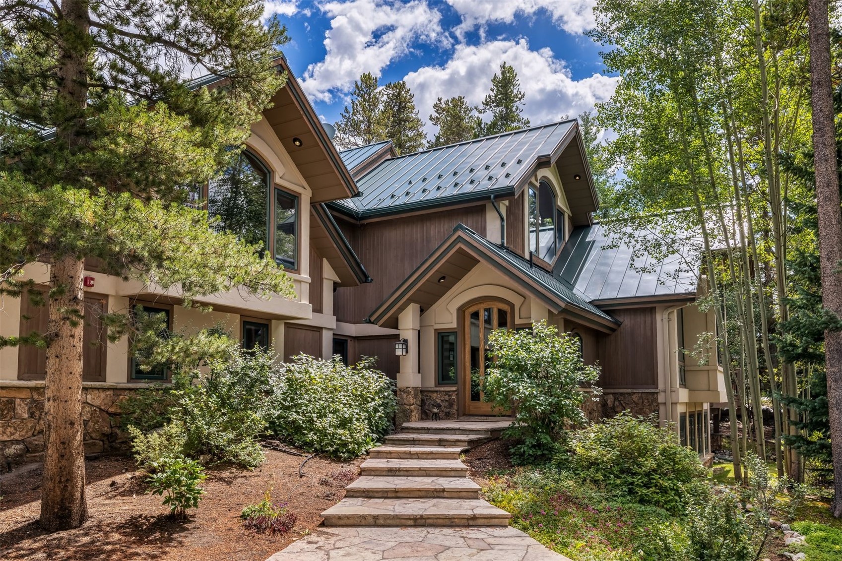 This mountain-style home features a striking exterior with a combination of dark wood siding, stone foundation accents, and a dark metal roof. A stone-paved walkway leads directly to the arched wooden front door, which is sheltered by a gabled portico. The surrounding landscape is lush with mature pine and aspen trees, creating a serene and secluded atmosphere.
