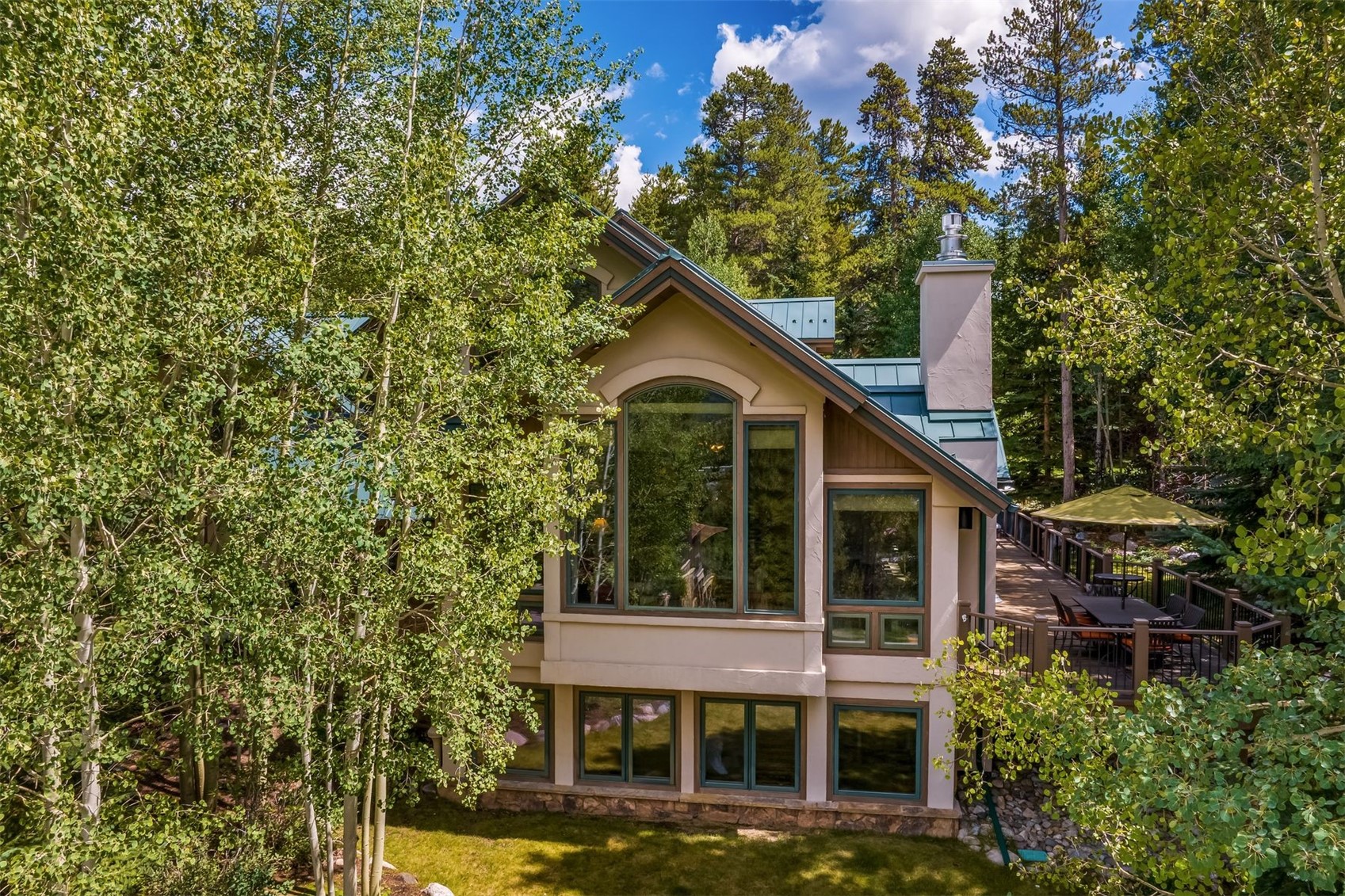 This elevated perspective captures the rear exterior of a multi-level mountain home nestled within a lush forest of aspen and pine trees. The structure features a prominent, tall arched window, a metal roof, and a spacious wooden deck equipped with outdoor dining furniture and a yellow umbrella. The lower level includes a row of windows overlooking a grassy lawn, creating a serene and secluded atmosphere.