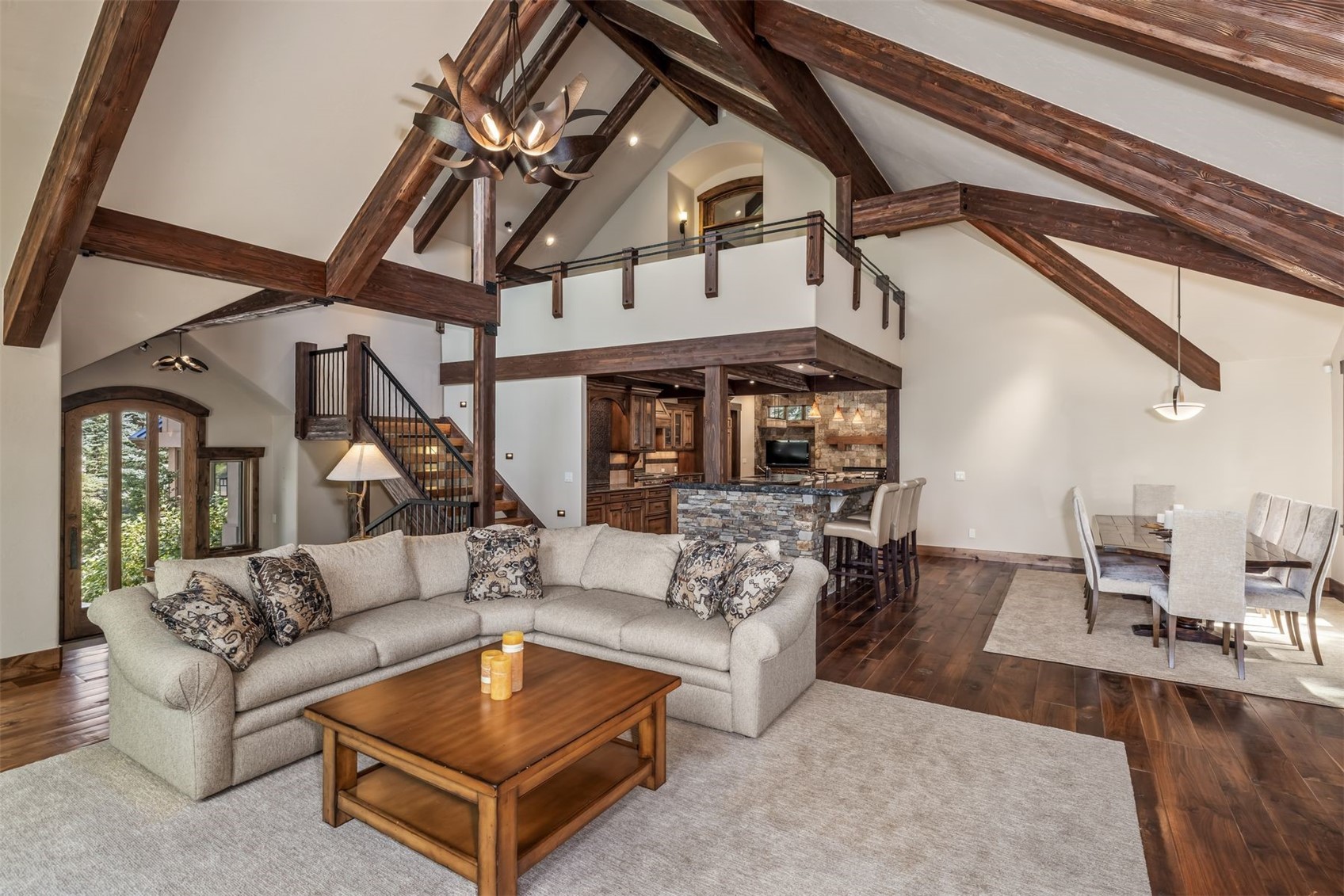 This expansive, open-concept living area features a dramatic vaulted ceiling with exposed dark wood beams and a rustic antler-style chandelier. The space includes a comfortable sectional sofa facing a wooden coffee table, with a stone-clad kitchen island and dining area visible in the background. A staircase with iron railings leads to an upper loft, creating a warm, lodge-inspired atmosphere with rich wood flooring and neutral-toned furnishings.