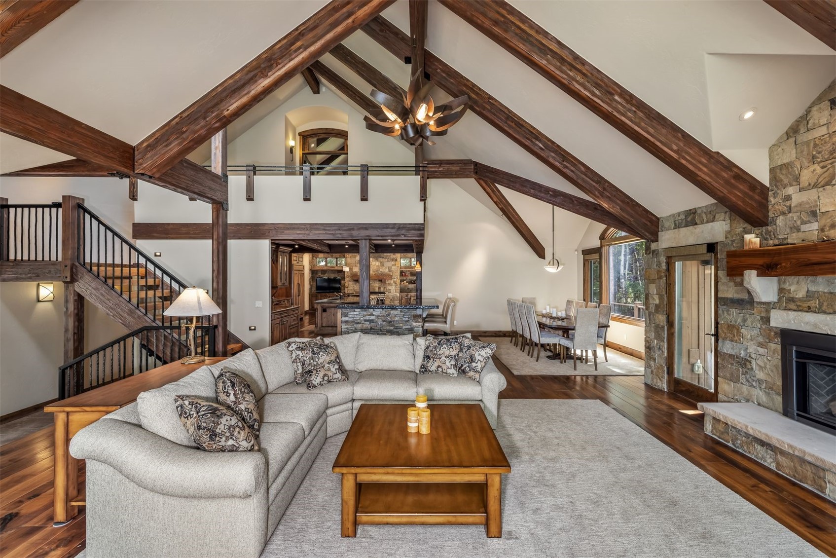 This expansive open-concept living area features a vaulted ceiling with exposed timber beams and a prominent stone-clad chimney breast. A large, neutral-toned sectional sofa faces a wooden coffee table, while a staircase with iron railings leads to an upper loft area. The space flows seamlessly into a dining area and kitchen, creating a warm, rustic atmosphere with rich wood flooring throughout.
