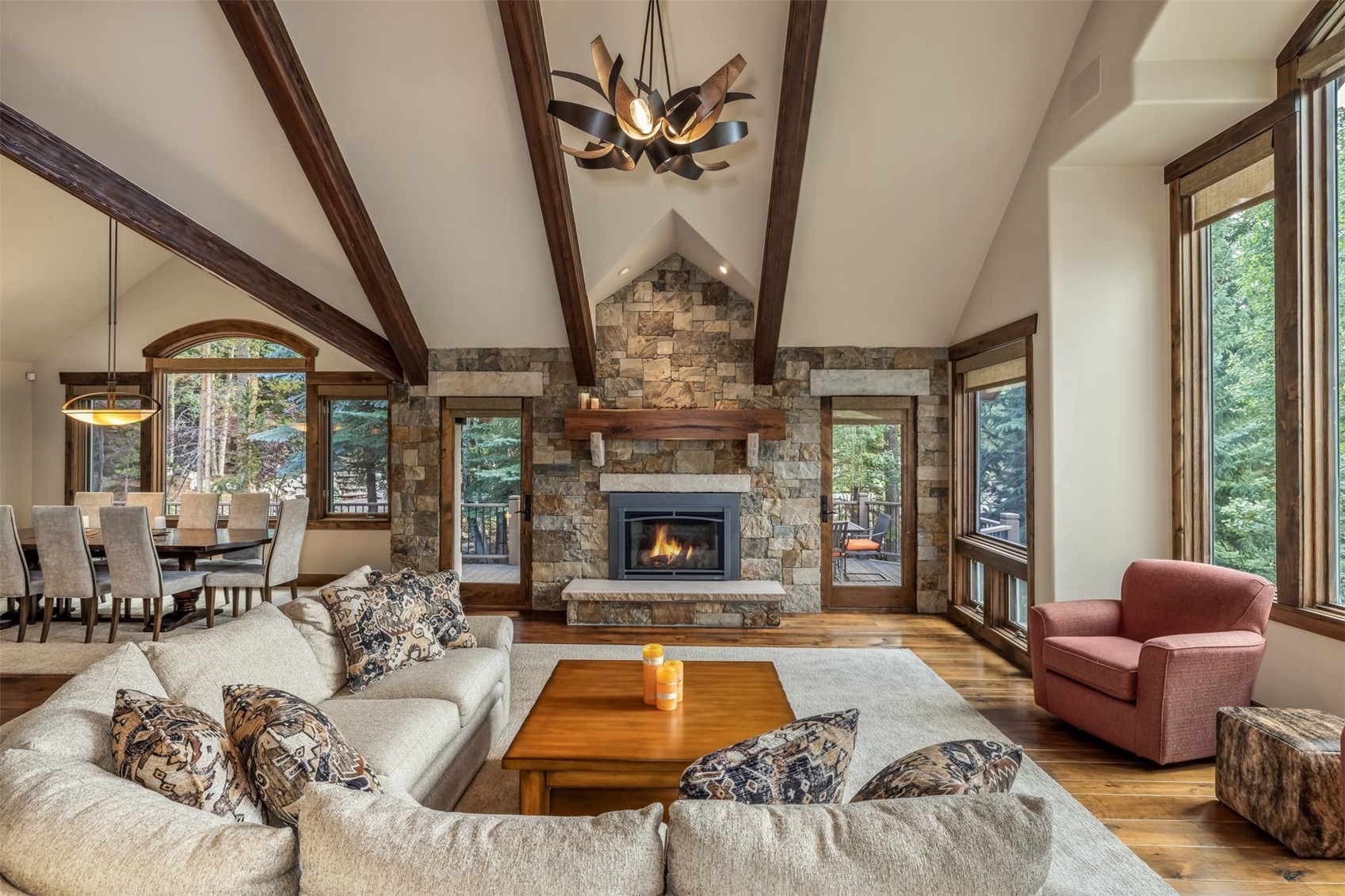 This spacious living room features a vaulted ceiling with exposed dark wood beams and a prominent stone-clad chimney breast centered between two glass doors. A large, light-colored sectional sofa faces a wooden coffee table, while a warm-toned armchair sits near tall windows that provide abundant natural light. The open-concept layout seamlessly connects to a dining area, creating an inviting atmosphere with a rustic, mountain-lodge aesthetic.