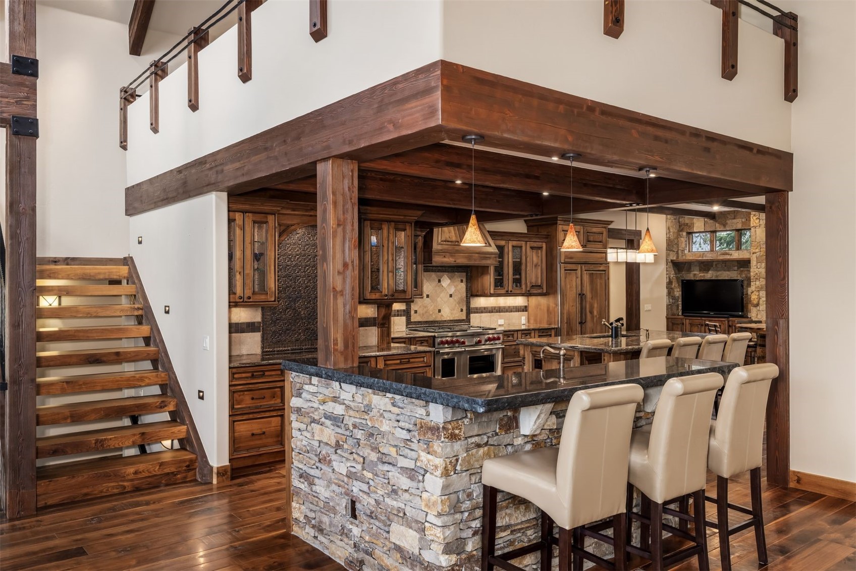 This rustic-style kitchen features a prominent stone-clad island with a dark countertop and three bar stools, serving as the focal point of the open-concept space. The area is framed by heavy, dark wood beams and includes custom cabinetry, a professional-grade range, and warm pendant lighting. To the left, a wooden staircase with an open design leads to an upper level, creating a cohesive, high-end mountain lodge aesthetic.