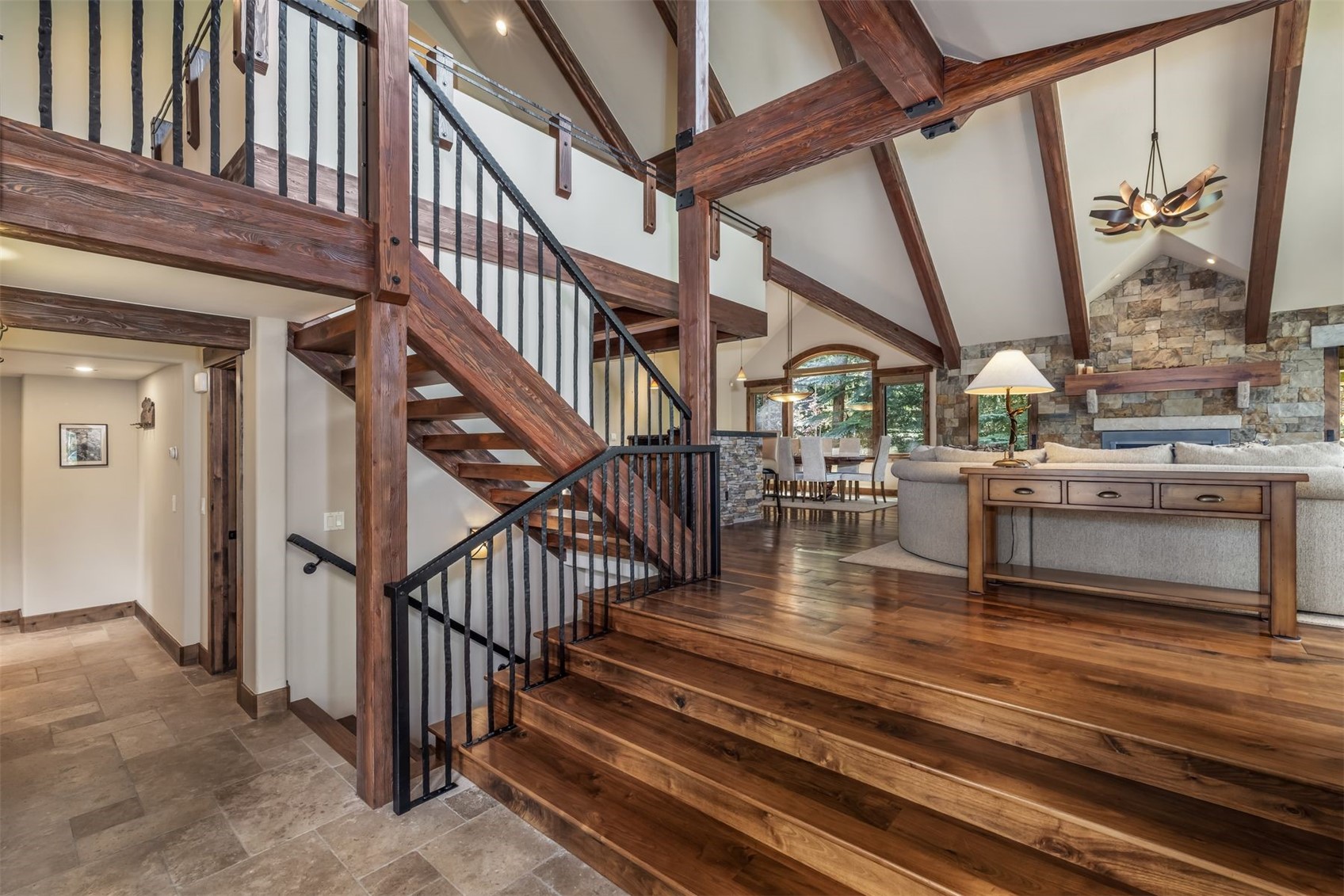 This grand entryway features a prominent wooden staircase with black metal railings that leads to an upper landing. The open-concept design flows into a spacious living area with vaulted ceilings, exposed timber beams, and a stone-clad chimney breast. The warm wood flooring and rustic architectural details create an inviting, lodge-inspired atmosphere.