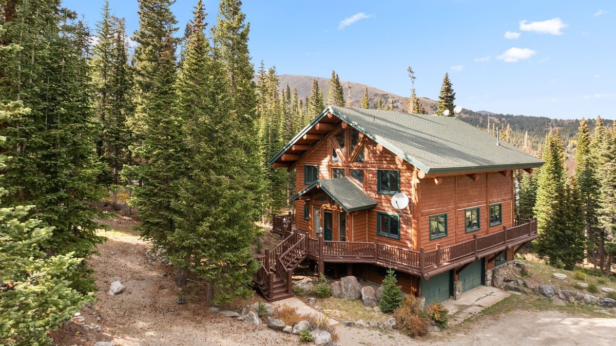 This elevated perspective captures a rustic log cabin nestled within a dense forest of pine trees. The home features a multi-level design with a wrap-around wooden deck, a green shingled roof, and a lower-level garage area. The natural wood exterior and mountainous backdrop create a serene, secluded atmosphere typical of a mountain retreat.