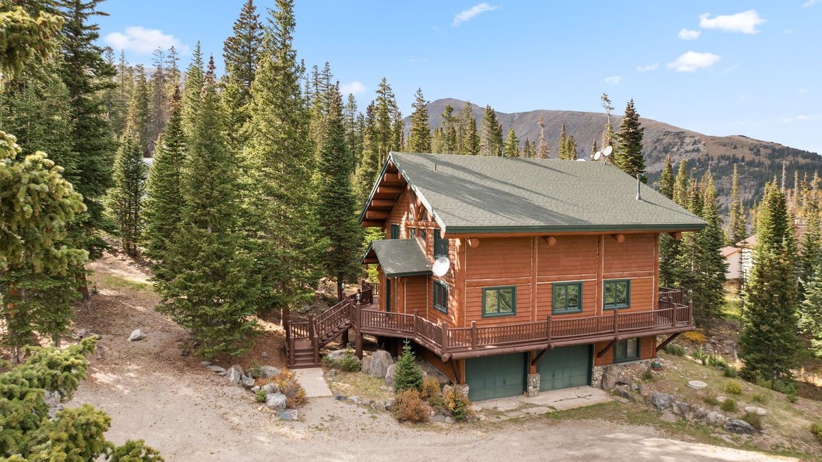 This rustic log cabin is nestled in a dense, mountainous forest setting, showcasing a multi-level design with a wrap-around wooden deck. The exterior features warm-toned horizontal logs, green-trimmed windows, and a two-car garage at the base level. The perspective is slightly elevated, capturing the home's integration into the rugged, pine-covered landscape under a clear blue sky.