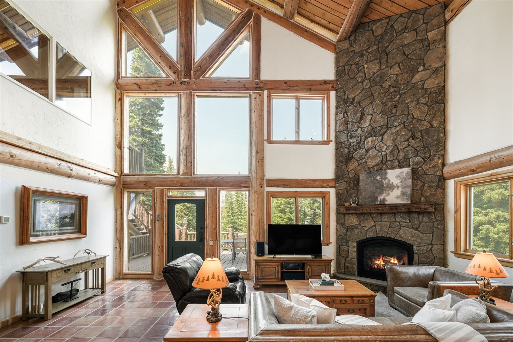 This spacious living room features a dramatic vaulted ceiling with exposed timber beams and a massive wall of windows that floods the space with natural light. A prominent stone-clad chimney breast serves as the focal point, complemented by warm-toned tile flooring and rustic log-style architectural accents. The room is furnished with comfortable leather seating and a wooden coffee table, creating a cozy, lodge-inspired atmosphere.