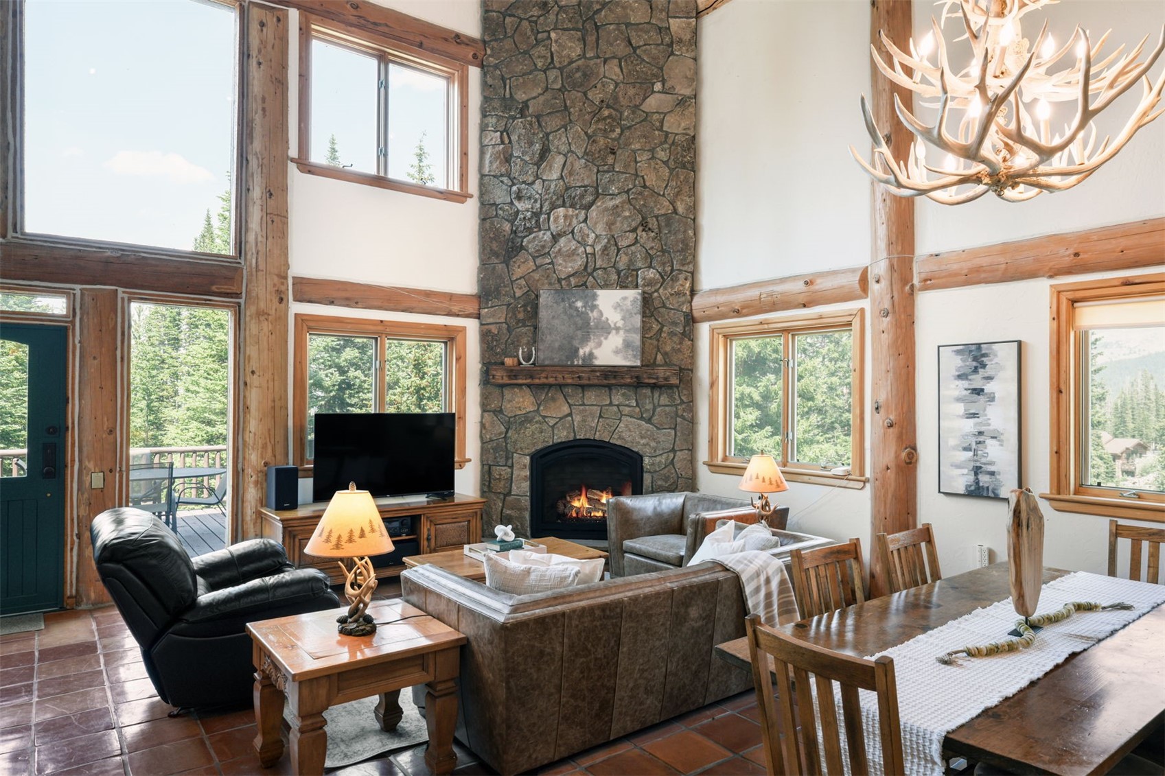 This rustic living room features a prominent stone-clad chimney breast that serves as the focal point of the space. The room is furnished with leather seating, a wooden coffee table, and a matching side table, all set against a backdrop of exposed timber beams and large windows that provide ample natural light. A distinctive antler chandelier hangs above, contributing to the cozy, mountain-lodge aesthetic of the open-concept living and dining area.