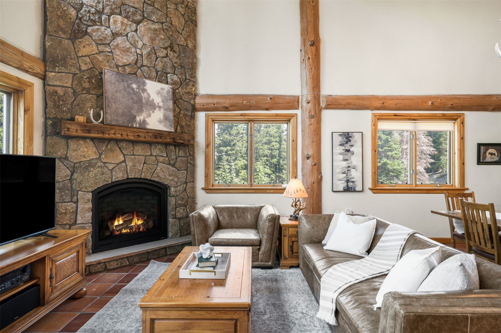 This cozy living room features a prominent stone-clad chimney breast with a built-in fireplace and a wooden mantel. The space is furnished with a leather sofa and armchair, complemented by a wooden coffee table and a neutral area rug. Exposed timber beams and warm wood-trimmed windows add to the rustic, lodge-inspired aesthetic of the room.