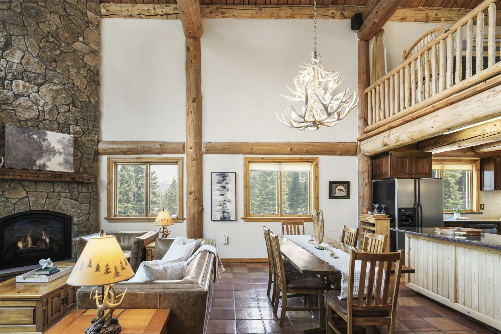 This open-concept living and dining area features a rustic mountain aesthetic with exposed log beams and a tall stone-clad chimney breast. The space includes a leather sofa, a wooden dining table with matching chairs, and a distinctive antler chandelier hanging from the high ceiling. Large windows provide natural light and views of the surrounding forest, while a wooden loft railing overlooks the main living area.