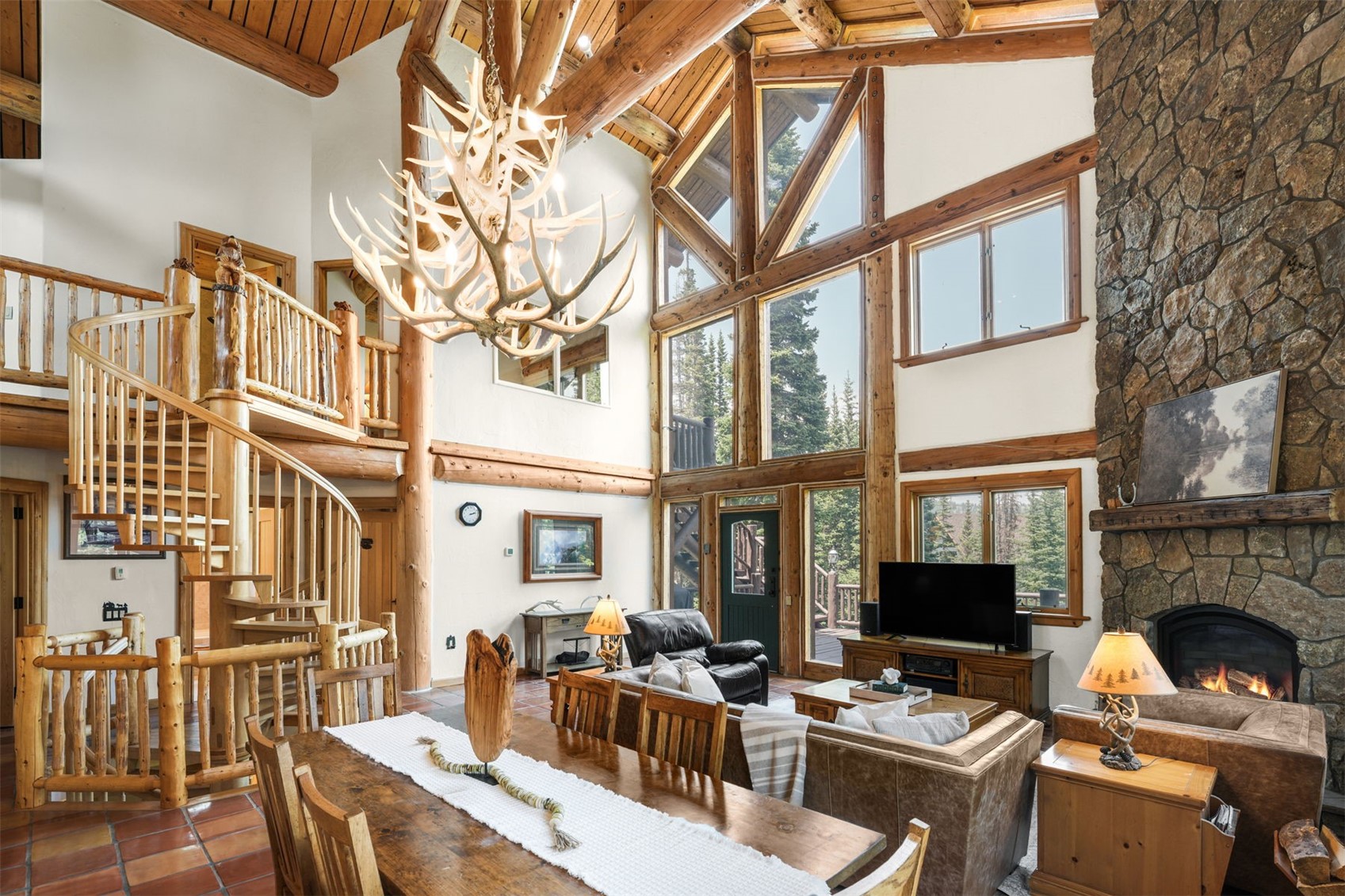 This expansive, open-concept living and dining area features a rustic lodge aesthetic with exposed log beams and a dramatic antler chandelier. A massive stone-clad chimney breast serves as a focal point, while large windows allow natural light to flood the space, highlighting the warm wood tones and tile flooring. The room is furnished with comfortable leather seating and a wooden dining table, creating a cozy yet grand atmosphere perfect for mountain living.