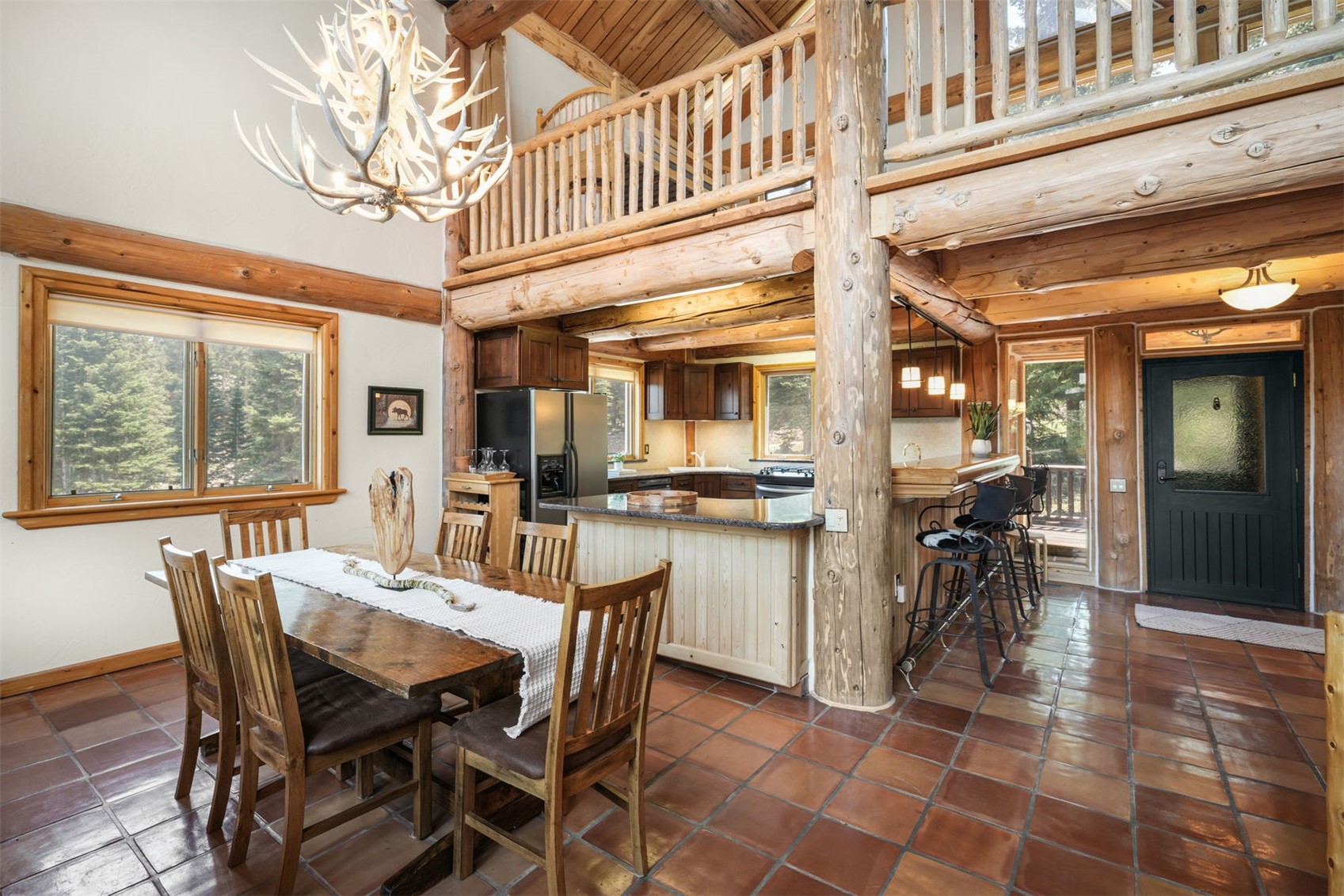 This rustic, open-concept kitchen and dining area features exposed log beams, a large antler chandelier, and warm-toned tile flooring. The kitchen includes a breakfast bar with stools, dark cabinetry, and a granite-topped island, while the adjacent dining space is anchored by a substantial wooden table. The perspective captures the cozy, lodge-style atmosphere, looking from the dining area toward the kitchen and a dark-framed exterior door.