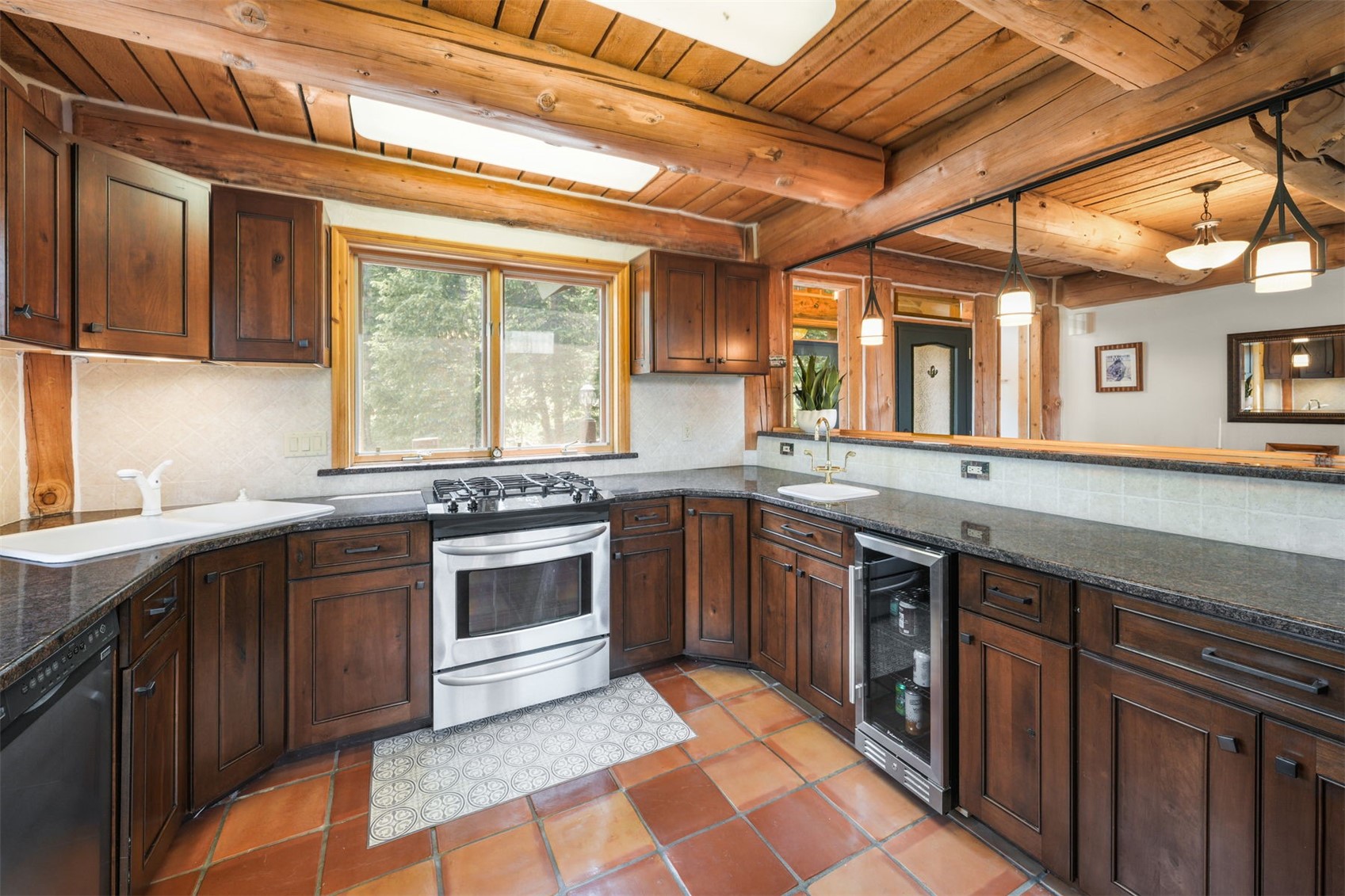 This rustic-style kitchen features warm wood cabinetry, dark stone countertops, and terracotta-toned floor tiles. The space is defined by exposed log ceiling beams and a pass-through window that connects to an adjacent room, creating an open and inviting atmosphere. Stainless steel appliances, including a gas range and a built-in beverage cooler, provide modern functionality within the traditional log-cabin aesthetic.