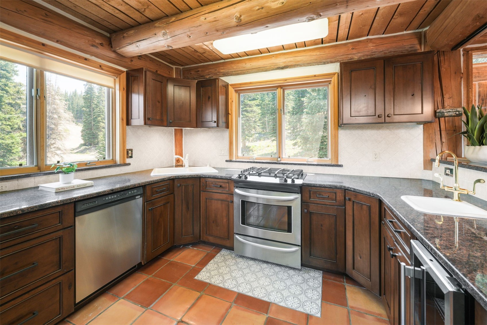 This rustic-style kitchen features warm wood cabinetry, dark stone countertops, and terracotta-toned floor tiles. The space is framed by exposed log ceiling beams and large windows that provide a scenic view of the surrounding forest. Stainless steel appliances, including a dishwasher and a gas range, are integrated into the U-shaped layout, creating a cozy and functional cooking environment.