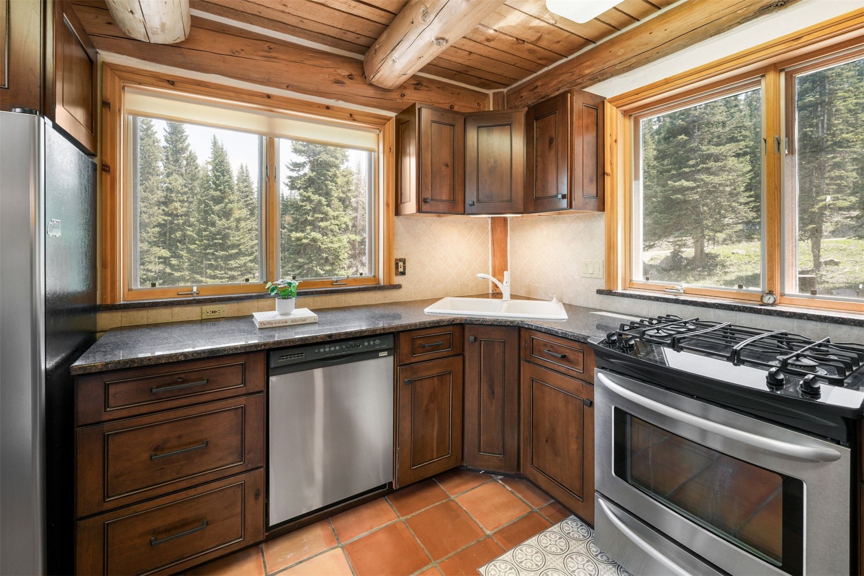 This rustic-style kitchen features warm wood cabinetry, dark stone countertops, and a corner sink setup. The space is framed by exposed log ceiling beams and large windows that provide a scenic view of the surrounding forest. The floor is finished with terracotta-colored tiles, and the kitchen is equipped with stainless steel appliances, including a dishwasher and a gas range.