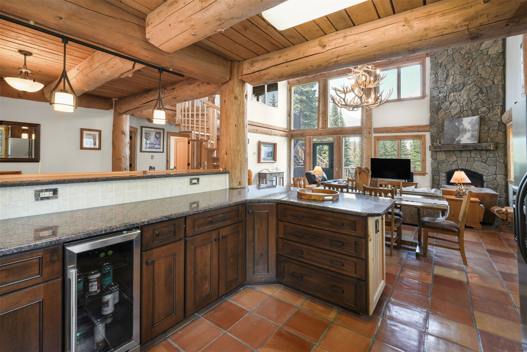 This rustic kitchen features dark wood cabinetry, a stone-topped island, and a built-in beverage cooler, seamlessly transitioning into an open-concept living and dining area. The space is defined by heavy timber beams, a stone-clad chimney breast, and large windows that provide an expansive view of the outdoors. The warm, earthy tones and natural textures create a cozy, lodge-like atmosphere perfect for mountain living.