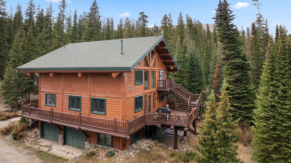 This elevated side view captures a rustic log cabin nestled within a dense evergreen forest. The home features a multi-level design with a wrap-around wooden deck, a two-car garage on the lower level, and a prominent staircase leading to the upper deck. The structure is characterized by its natural wood siding, dark-trimmed windows, and a pitched roof, blending seamlessly into the mountainous, wooded landscape.