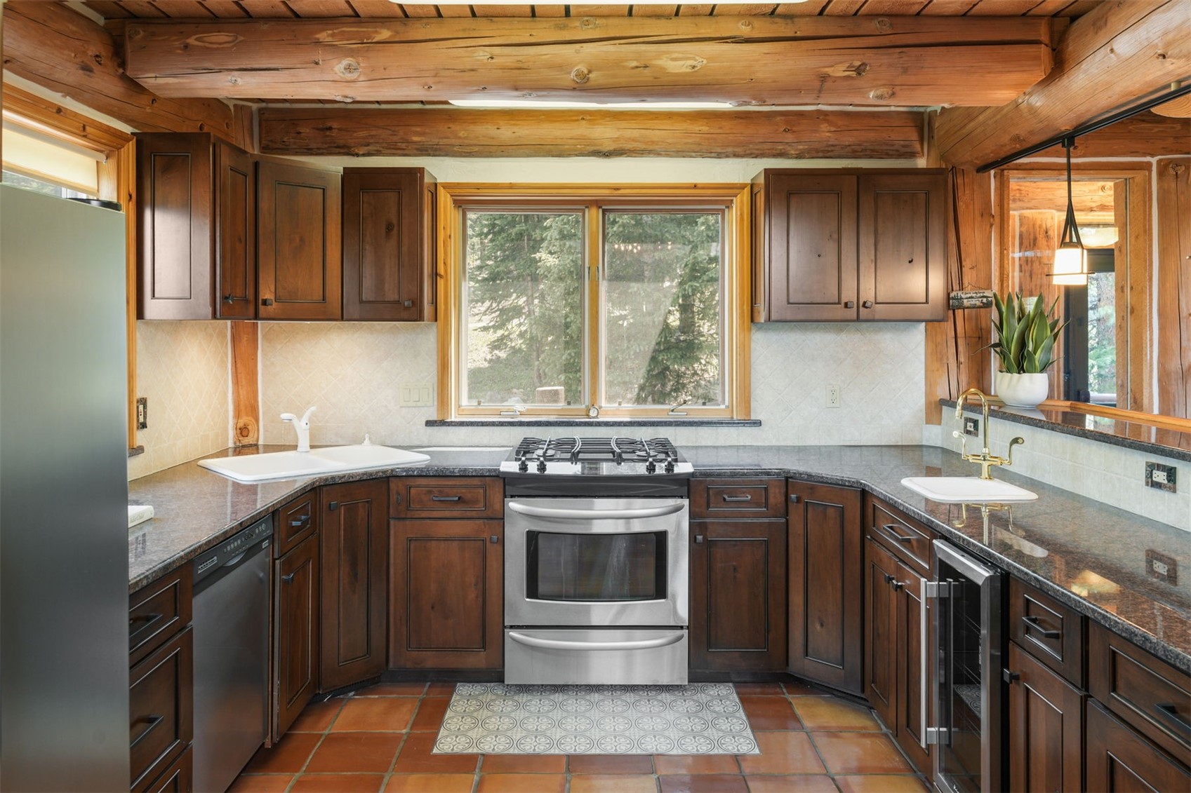 This rustic-style kitchen features dark wood cabinetry, granite countertops, and a central stainless steel gas range. The space is framed by exposed log ceiling beams and terracotta-toned floor tiles, creating a warm, cabin-like atmosphere. A large window above the stove provides natural light, while a secondary sink and a wine cooler are integrated into the perimeter layout.