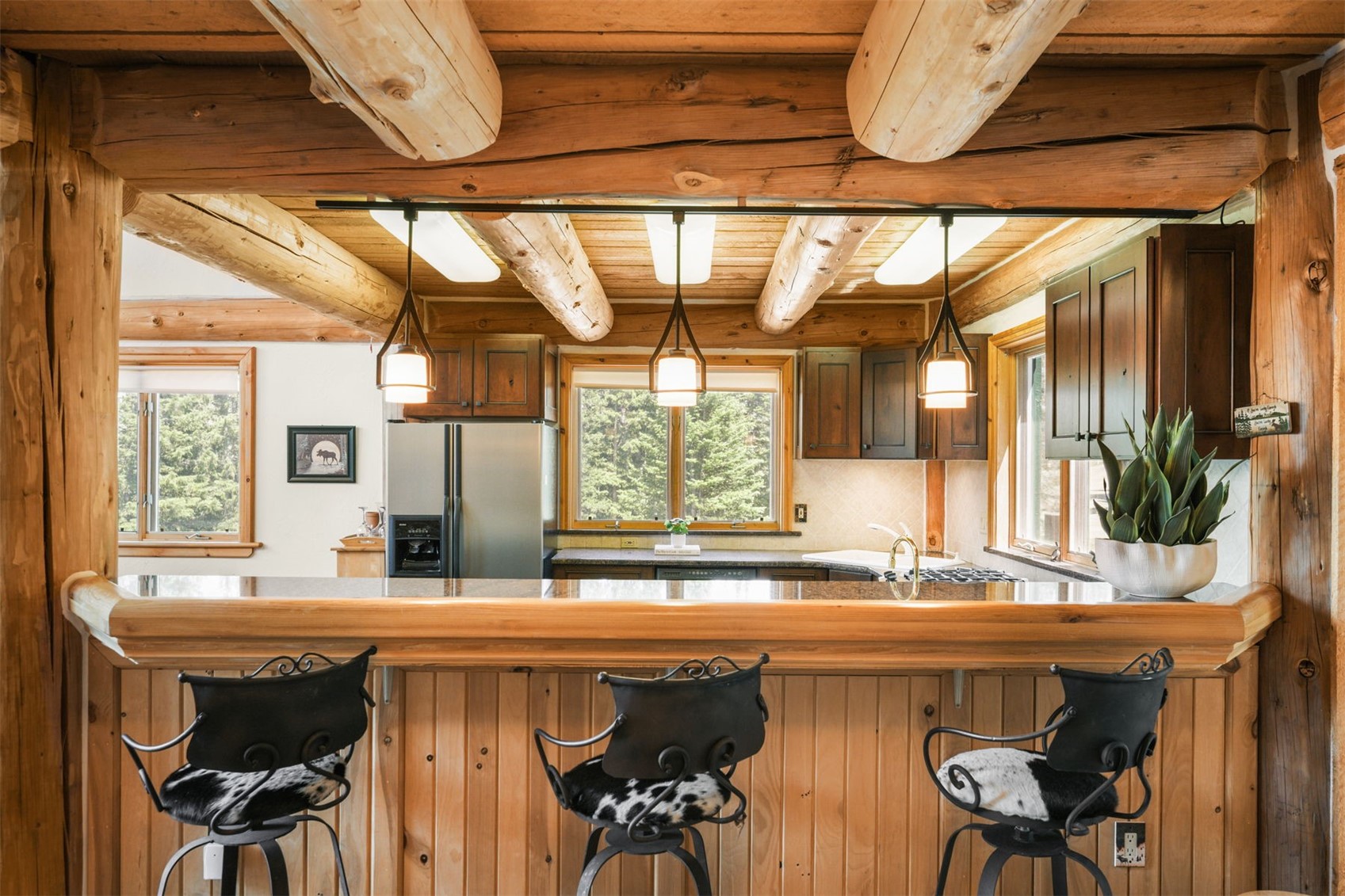 This rustic kitchen features a prominent breakfast bar with three stools upholstered in cowhide, set against a backdrop of exposed log beams and wood-paneled walls. The space includes stainless steel appliances, dark cabinetry, and a granite countertop, all illuminated by three pendant lights hanging from a track system. The warm, natural wood tones and cabin-style architecture create a cozy, inviting atmosphere with a view through the window into the surrounding forest.