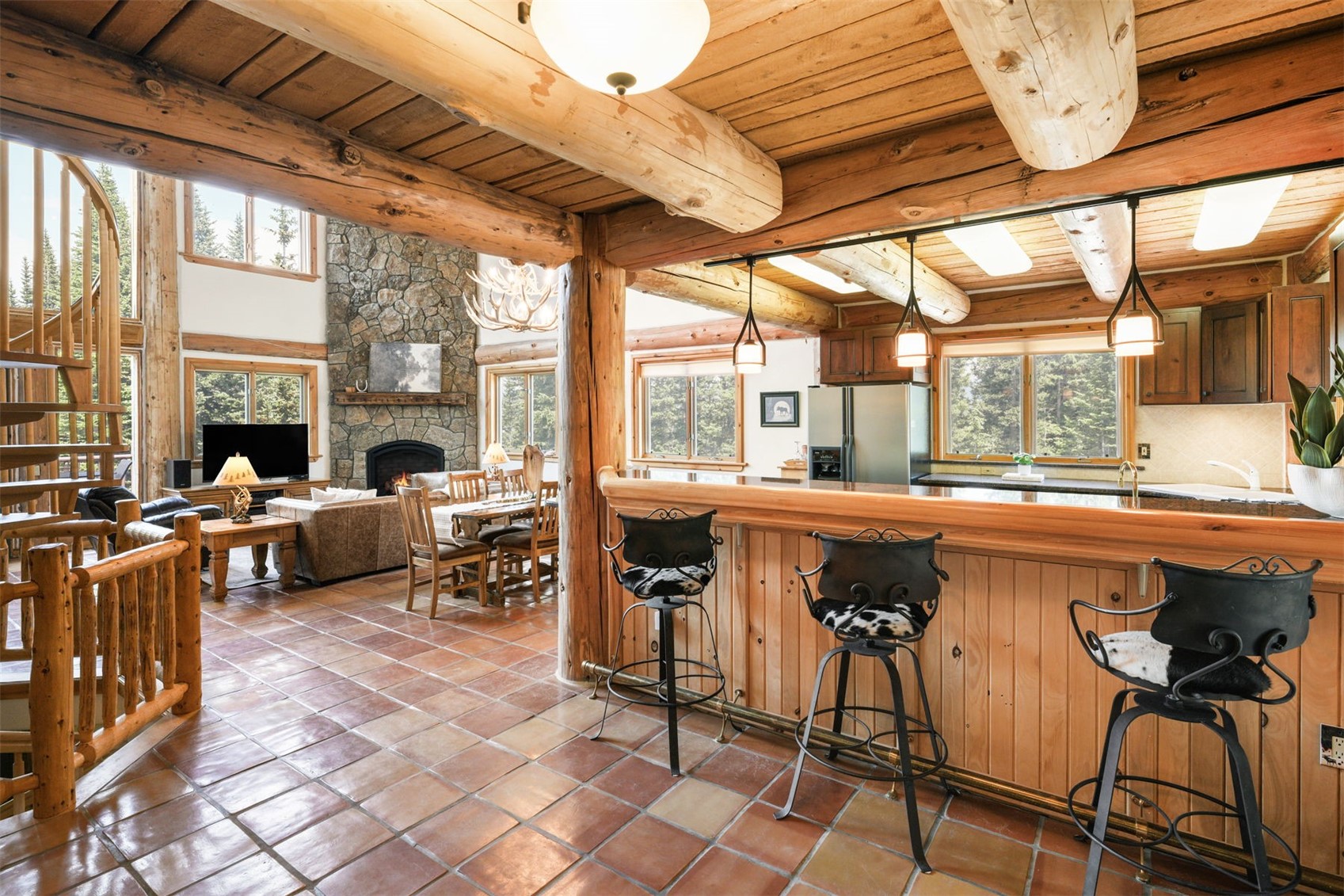 This rustic, open-concept living space features a prominent log-beam ceiling and warm-toned tile flooring throughout. The foreground showcases a wooden breakfast bar with three metal stools, while the background reveals a cozy living area centered around a stone-clad chimney breast and a dining space illuminated by a decorative antler chandelier. The perspective is captured from the kitchen area, looking across the open floor plan toward the living room and spiral staircase.