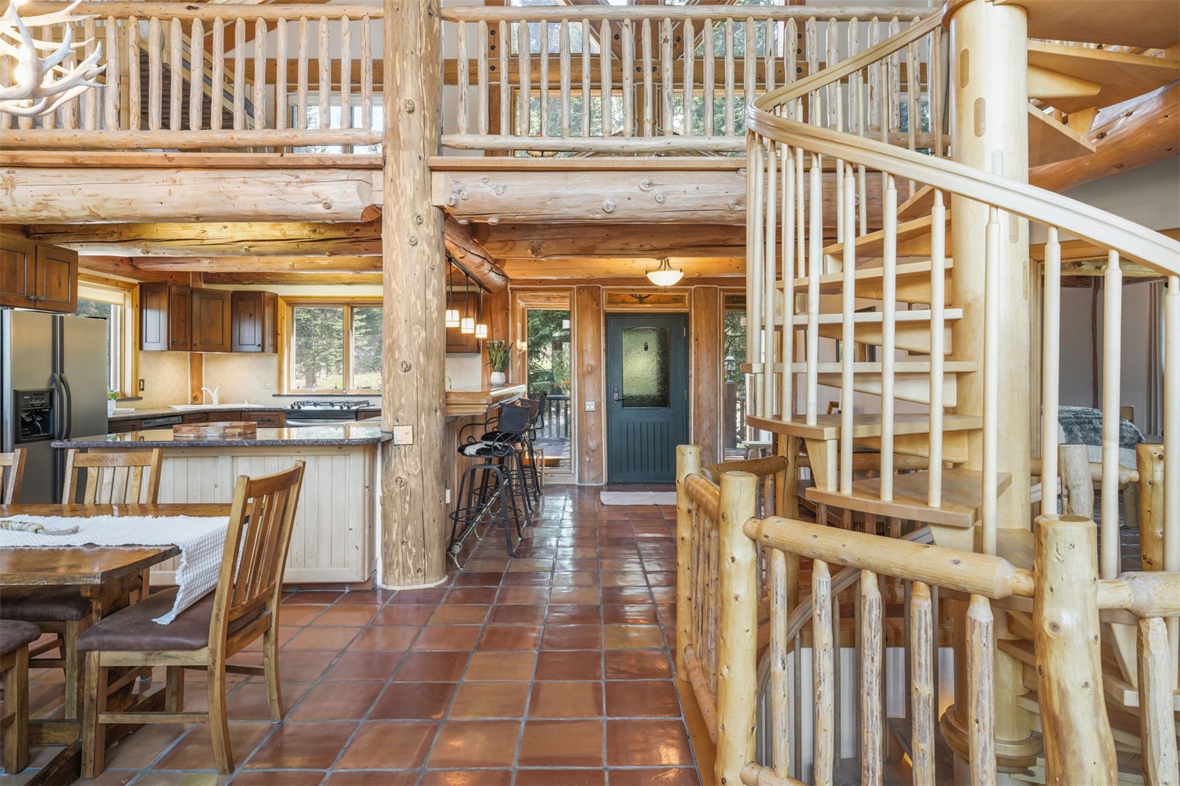 This rustic, open-concept interior features a log-cabin aesthetic with exposed timber beams and a prominent spiral staircase. The kitchen area includes dark wood cabinetry, a stone-topped island with bar seating, and a dining table in the foreground, all set against a backdrop of warm-toned tile flooring. The perspective captures the transition from the dining and kitchen space toward the main entryway, emphasizing the home's spacious, multi-level design.