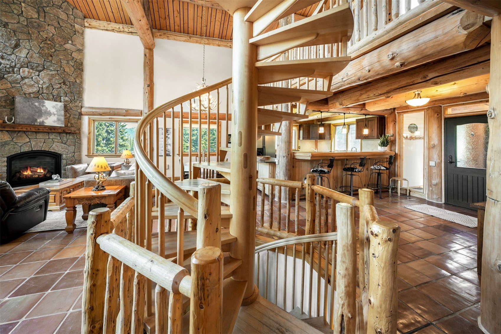 This rustic interior features a prominent spiral staircase crafted from light wood, serving as a central architectural focal point. To the left, a stone-clad chimney breast rises above a fireplace in the living area, while the background reveals an open-concept kitchen with a breakfast bar and stools. The space is characterized by exposed log beams, warm wood tones, and terracotta-style tile flooring, creating a cozy and inviting mountain lodge aesthetic.