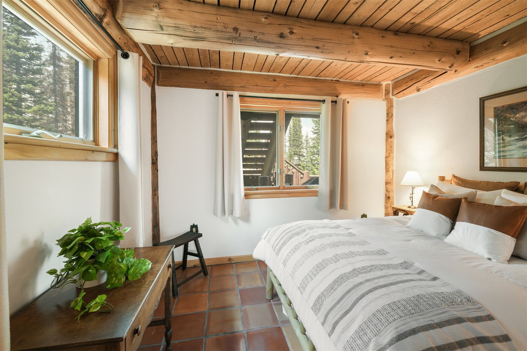 This cozy bedroom features a rustic aesthetic with exposed wooden ceiling beams and warm-toned terracotta floor tiles. A comfortable bed with layered bedding and brown accent pillows serves as the focal point, complemented by a wooden desk and a small stool. Large windows allow natural light to fill the space, offering a view of the surrounding forest and an exterior staircase.
