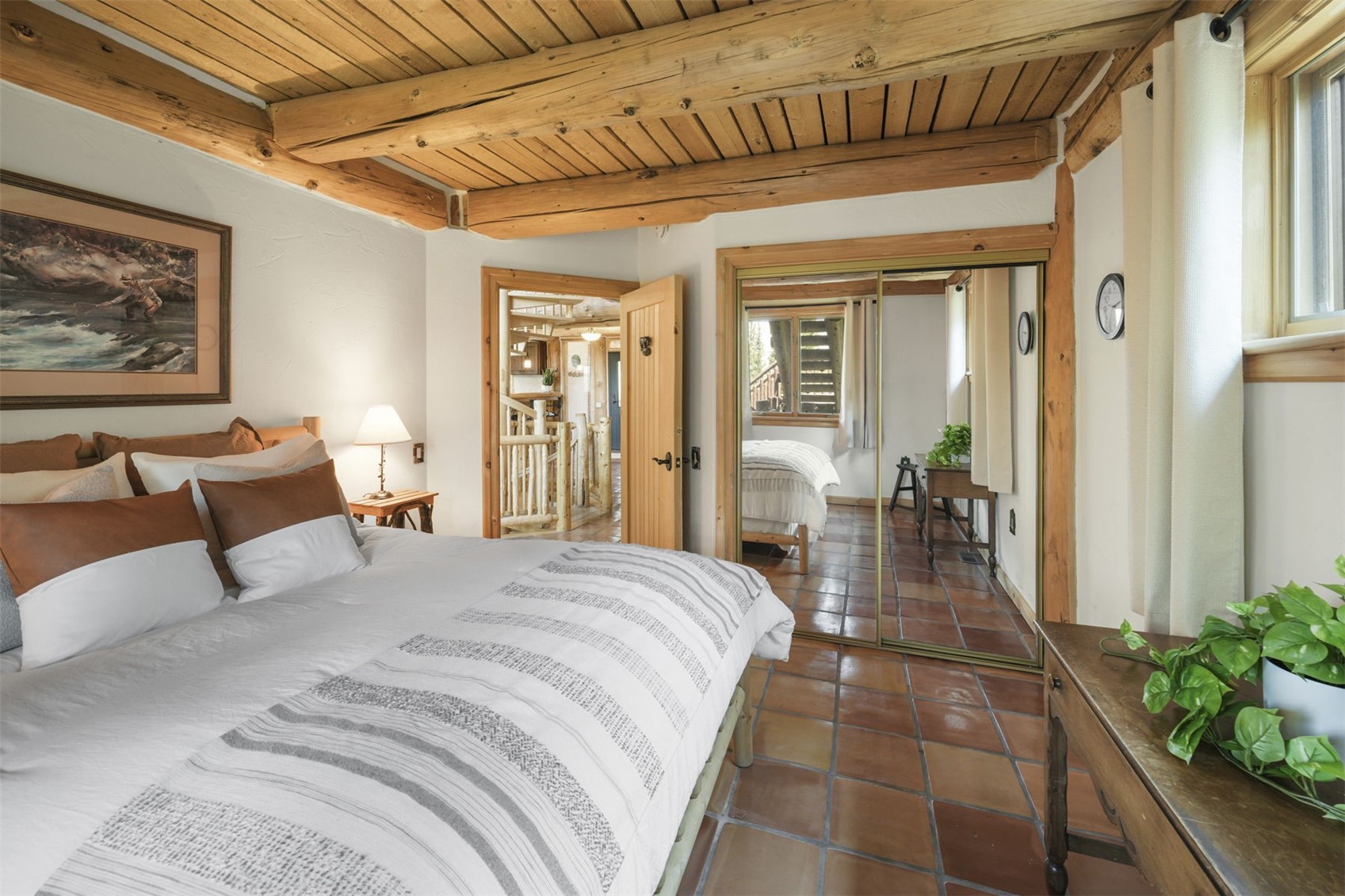 This cozy bedroom features a rustic aesthetic with exposed log ceiling beams and warm-toned terracotta tile flooring. A large bed with layered bedding serves as the focal point, while a mirrored closet door reflects the room and provides a view into the adjacent hallway. The space is accented by a wooden side table, a framed landscape print, and a small desk with a potted plant, creating a comfortable and inviting atmosphere.