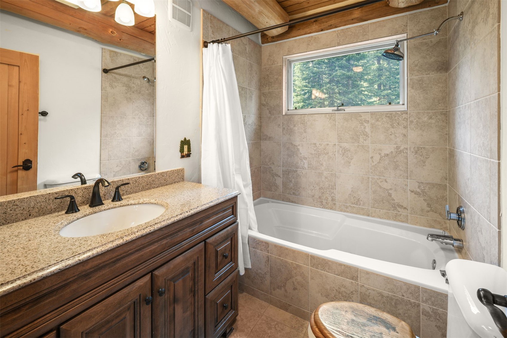 This rustic-style bathroom features a dark wood vanity topped with a speckled stone countertop and a white undermount sink. A tiled shower-tub combination is positioned beneath a rectangular window that looks out onto a wooded area, while exposed wooden ceiling beams add a warm, cabin-like aesthetic. The room is completed by a mirror above the vanity and a small wooden stool in the foreground.