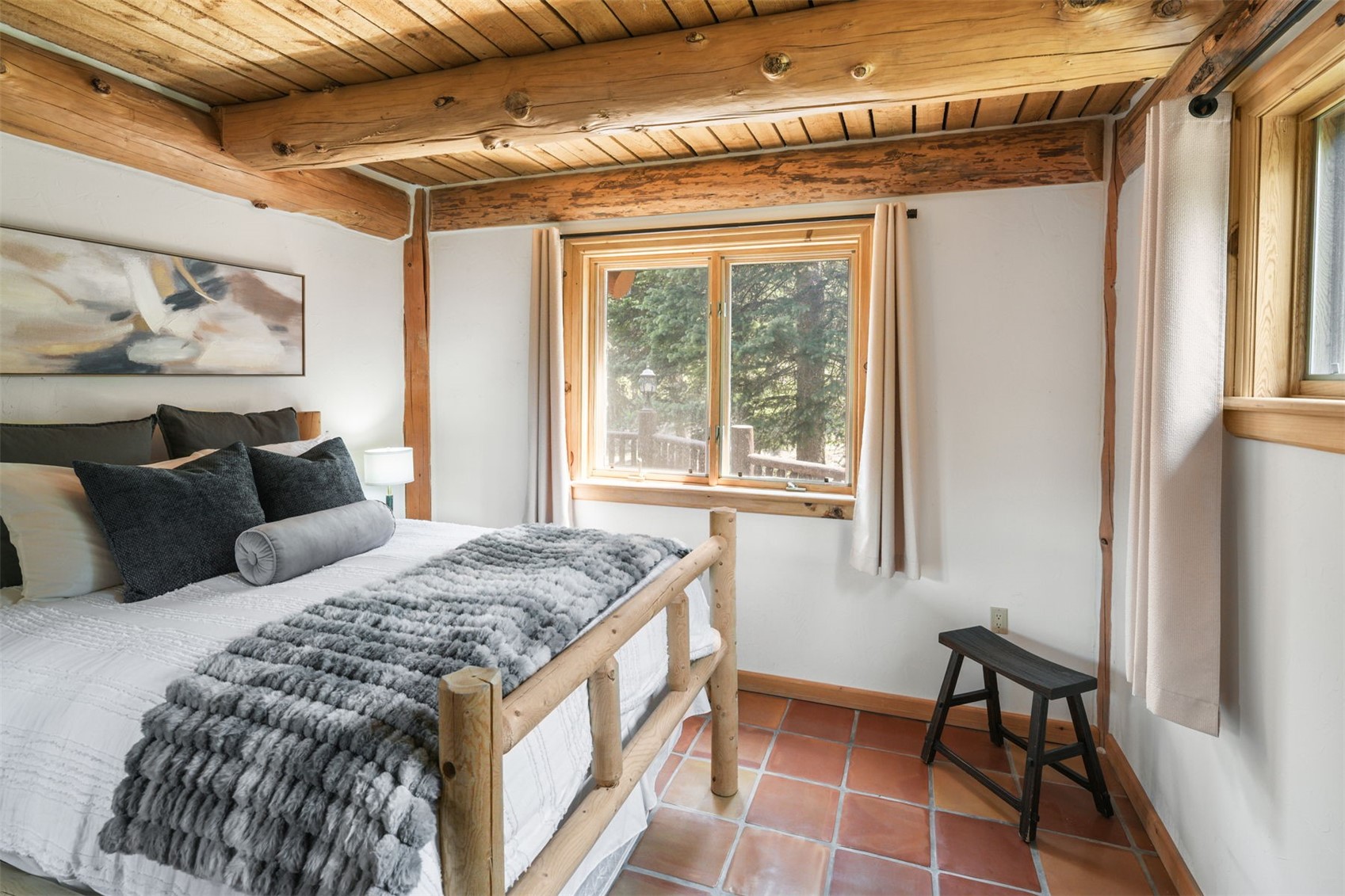 This cozy bedroom features a rustic aesthetic with exposed log ceiling beams and terracotta-style floor tiles. The room is furnished with a log-frame bed topped with a textured gray throw and plush pillows, complemented by a small black wooden bench. A window with light-colored curtains provides a view of the surrounding trees, creating a warm and inviting atmosphere.