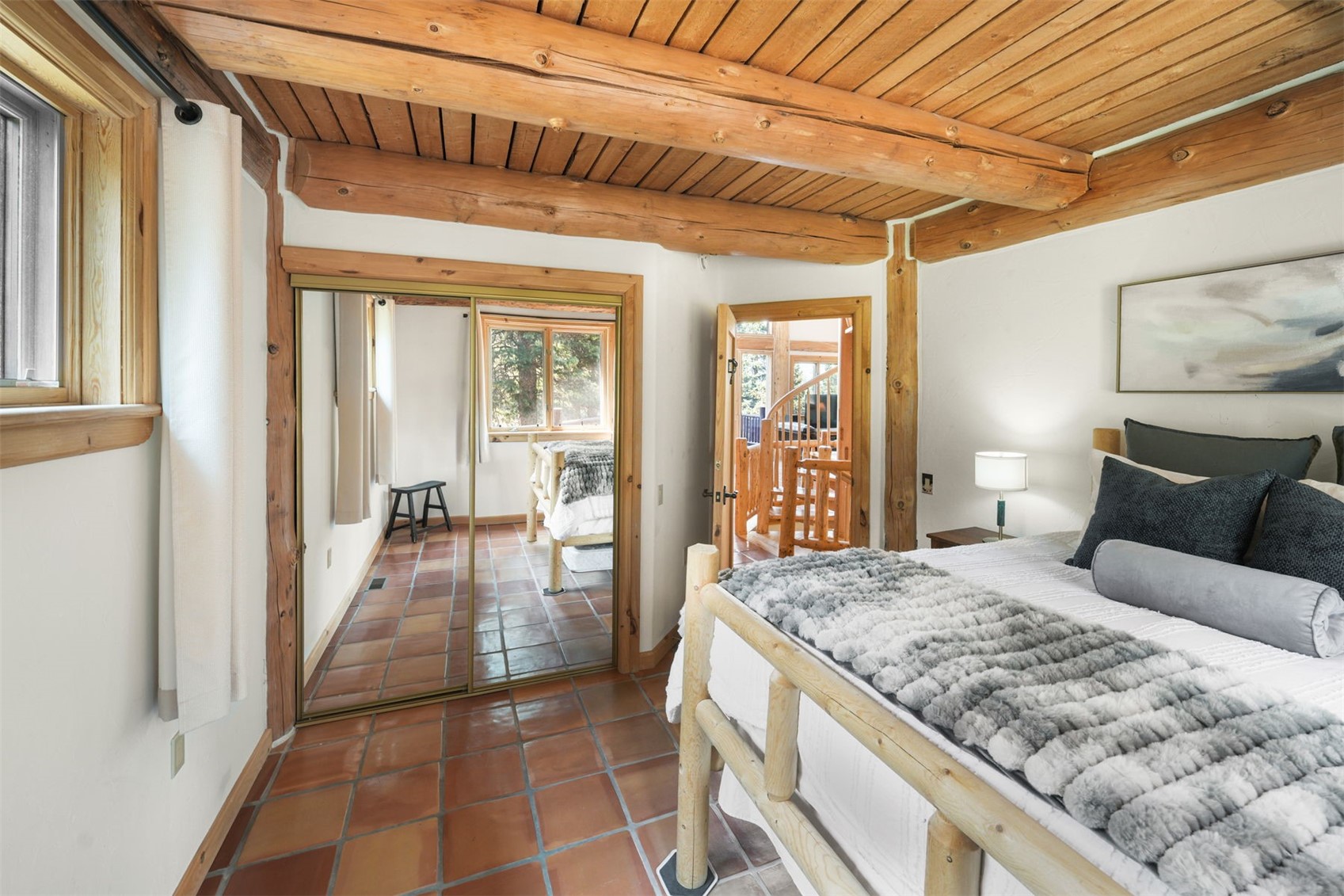 This rustic-style bedroom features exposed log ceiling beams and warm-toned terracotta tile flooring, creating a cozy, cabin-like atmosphere. A log-frame bed with a plush, textured grey throw blanket serves as the focal point, while a large mirrored closet door reflects the room's interior and adds a sense of depth. The space includes a doorway leading to a staircase, emphasizing the home's unique architectural character and connection to other levels.