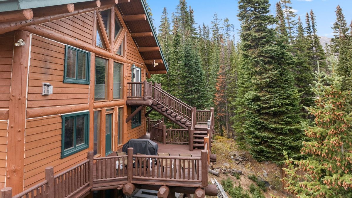 This side view of a rustic log cabin showcases a multi-level wooden deck and an exterior staircase leading to an upper balcony. The structure features warm-toned horizontal log siding, dark-framed windows, and a prominent gable roofline. Surrounded by a dense forest of evergreen trees, the home offers a secluded, mountain-inspired aesthetic.