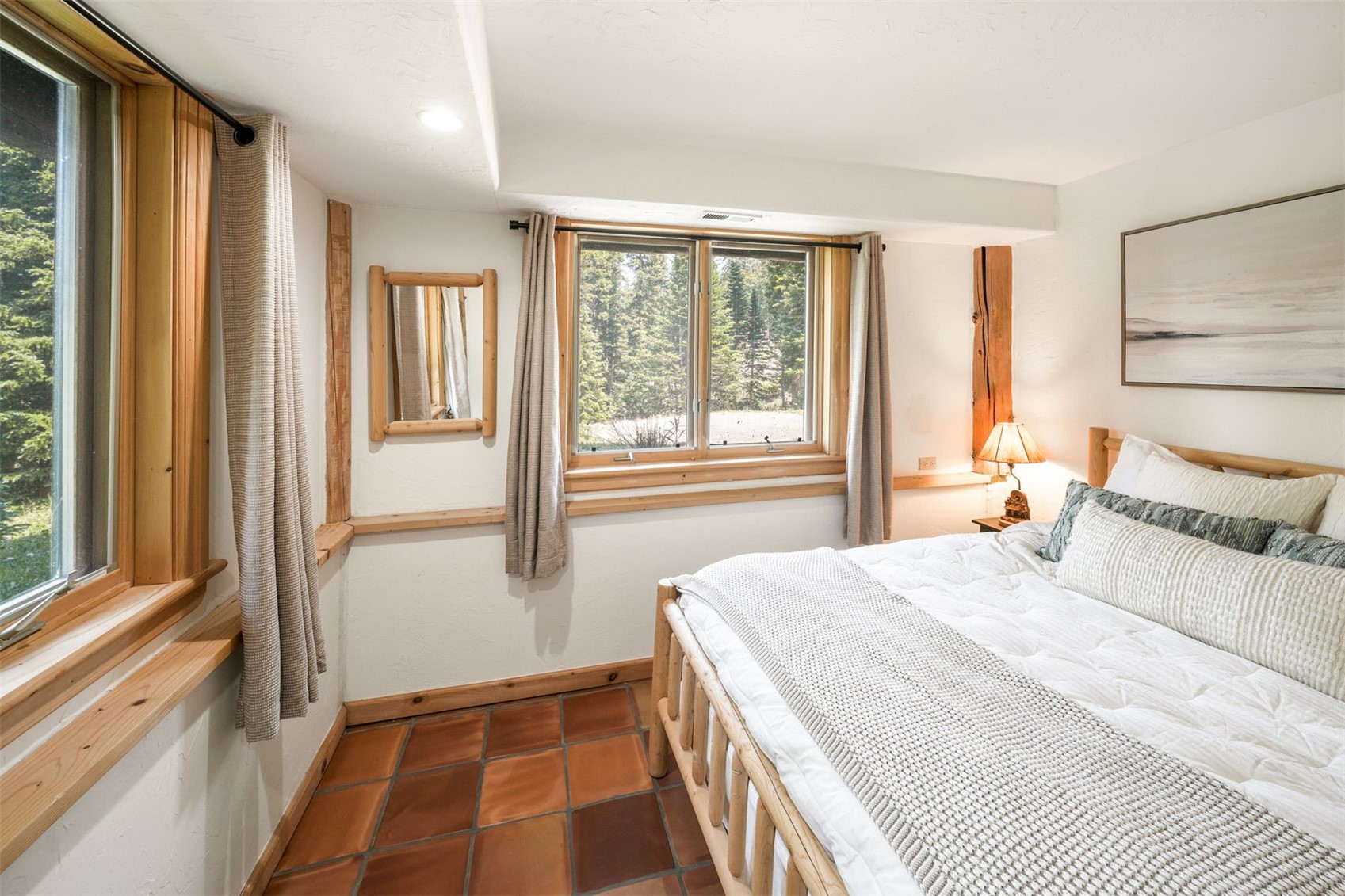 This cozy bedroom features a rustic aesthetic with light wood trim, a log-style bed frame, and warm-toned square floor tiles. Large windows provide a clear view of the surrounding forest, while a small mirror and a bedside lamp add functional charm to the space. The room is decorated with neutral-toned bedding and a framed landscape print, creating a serene and inviting atmosphere.