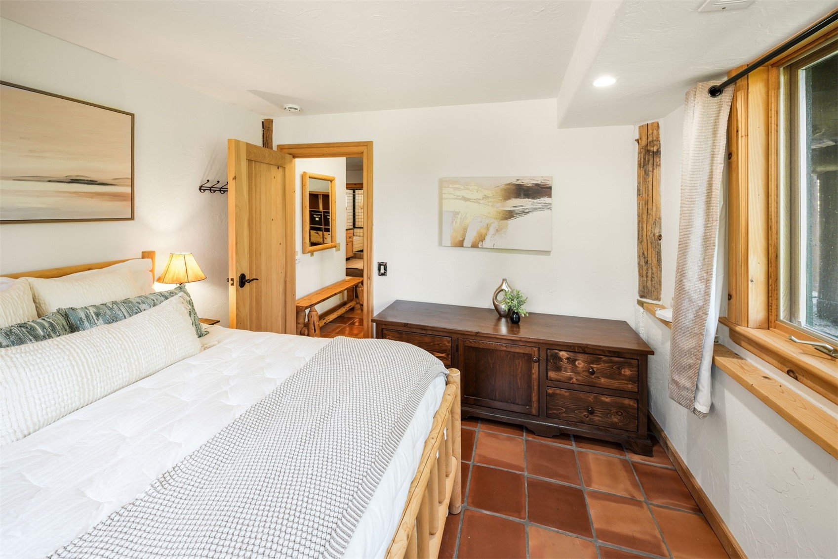 This cozy bedroom features a rustic aesthetic with a log-style bed frame and warm-toned terracotta tile flooring. A dark wood dresser sits against the wall beneath abstract artwork, while a doorway leads to an adjacent hallway with a wooden bench. The room is illuminated by a bedside lamp and natural light streaming through a window framed by light wood trim.