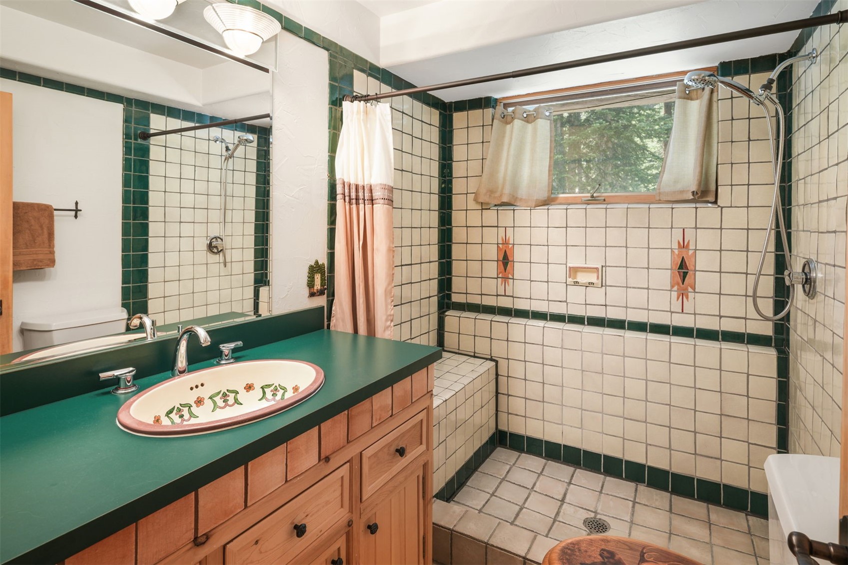 This bathroom features a distinctive Southwestern-inspired design with cream-colored square tiles accented by dark green trim and decorative geometric motifs. The vanity includes a light wood cabinet topped with a dark green counter and a decorative floral-patterned sink basin. A walk-in shower area with a built-in bench and a handheld showerhead is positioned next to a window, creating a unique and rustic aesthetic.