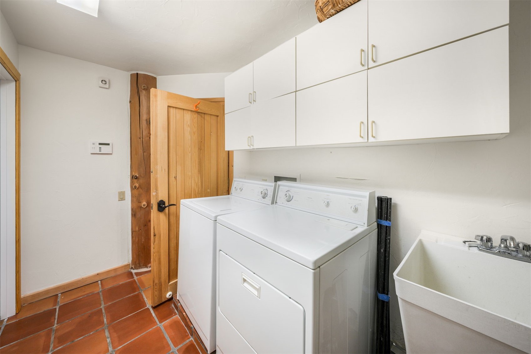 This functional laundry room features terracotta-style floor tiles and white cabinetry providing ample storage space above the washer and dryer. A utility sink is positioned to the right, while a rustic wooden door with a vertical plank design adds a touch of character to the space. The room is brightly lit, creating a clean and practical environment for household chores.